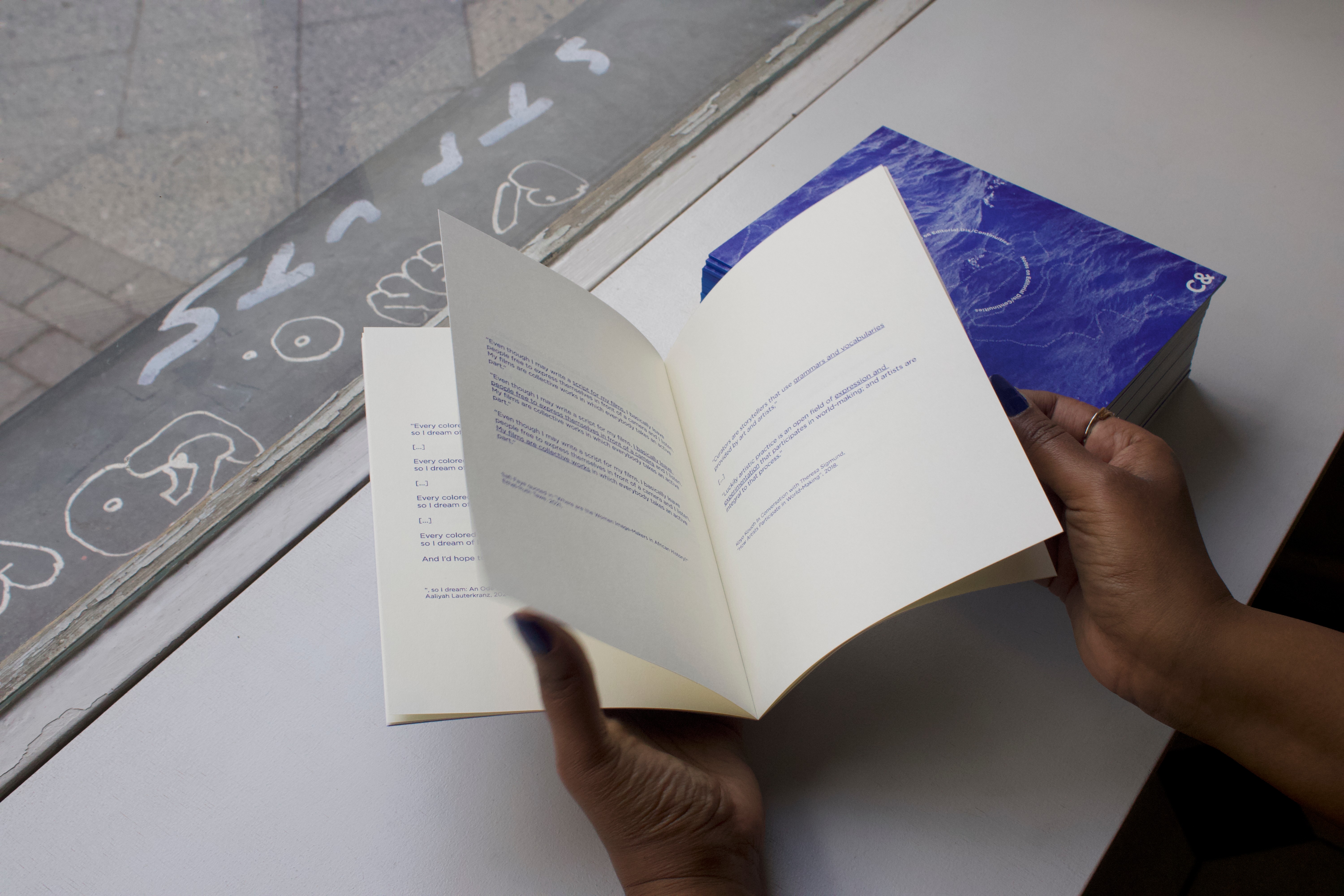 Hands holding an open publication with text, next to a stack of blue books, with graffiti visible through a window in the background.
