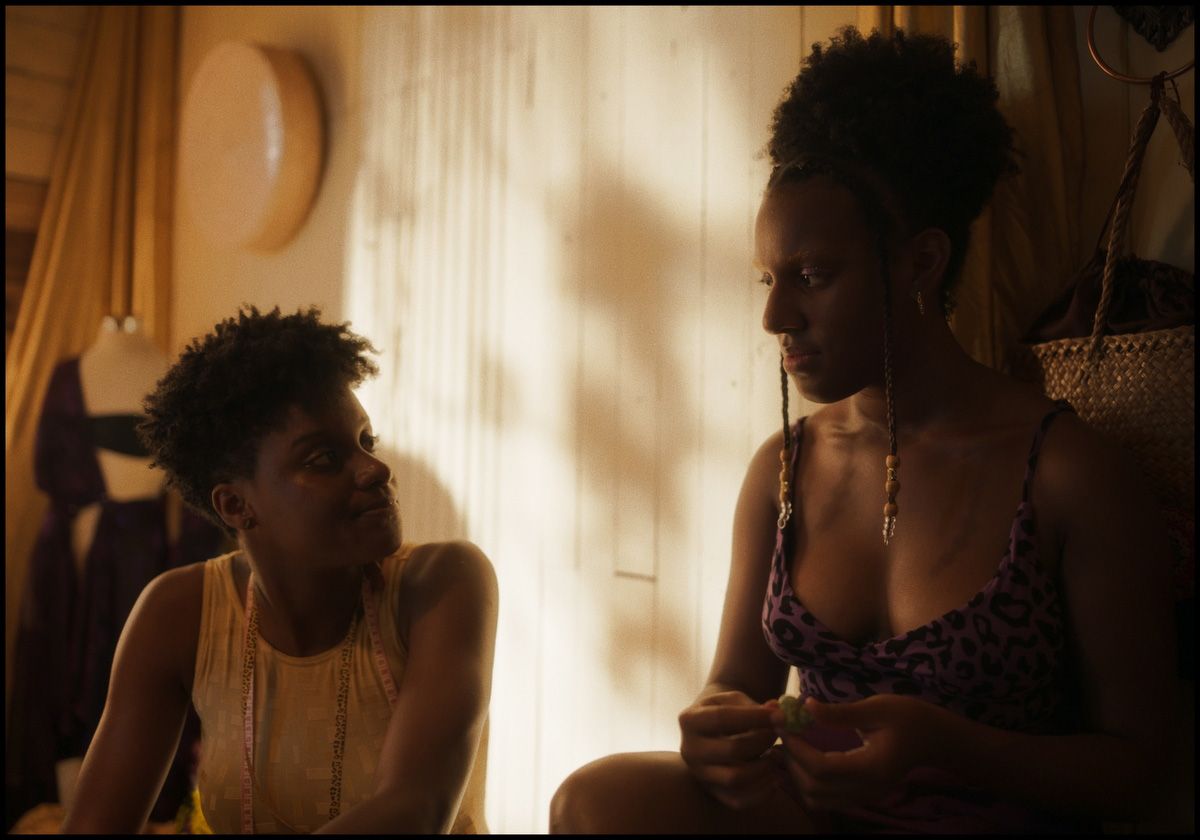 Two Black women in warm light, one with a measuring tape looking at the other with beaded braids holding a green object.