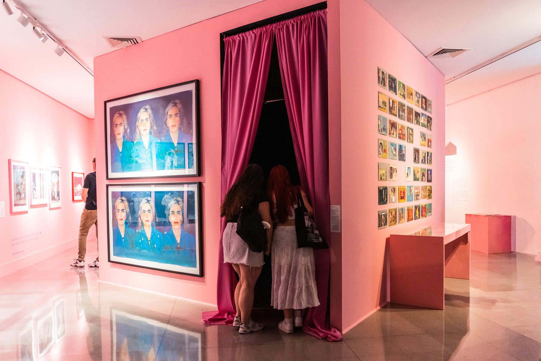 A pink art gallery with pop art portraits and visitors looking into a pink-curtained doorway.
