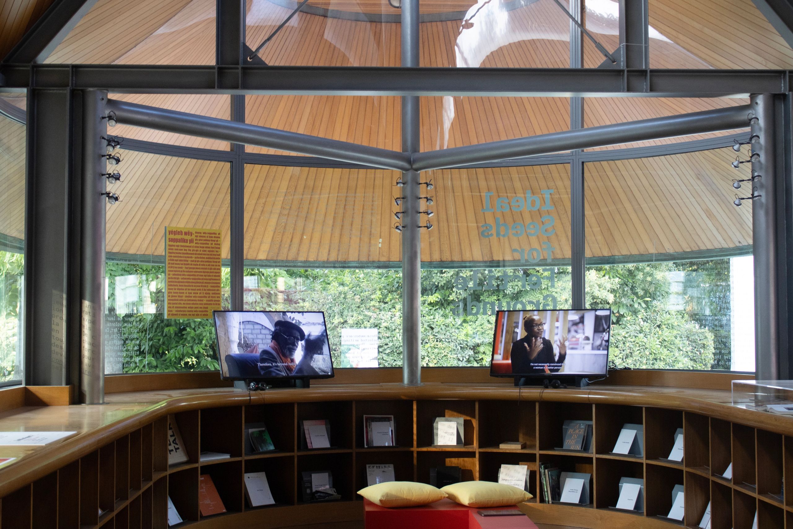 A modern, circular room with a curved wooden counter displaying books and two video screens. Large windows offer a view of trees, and the ceiling features wooden panels and exposed metal beams.