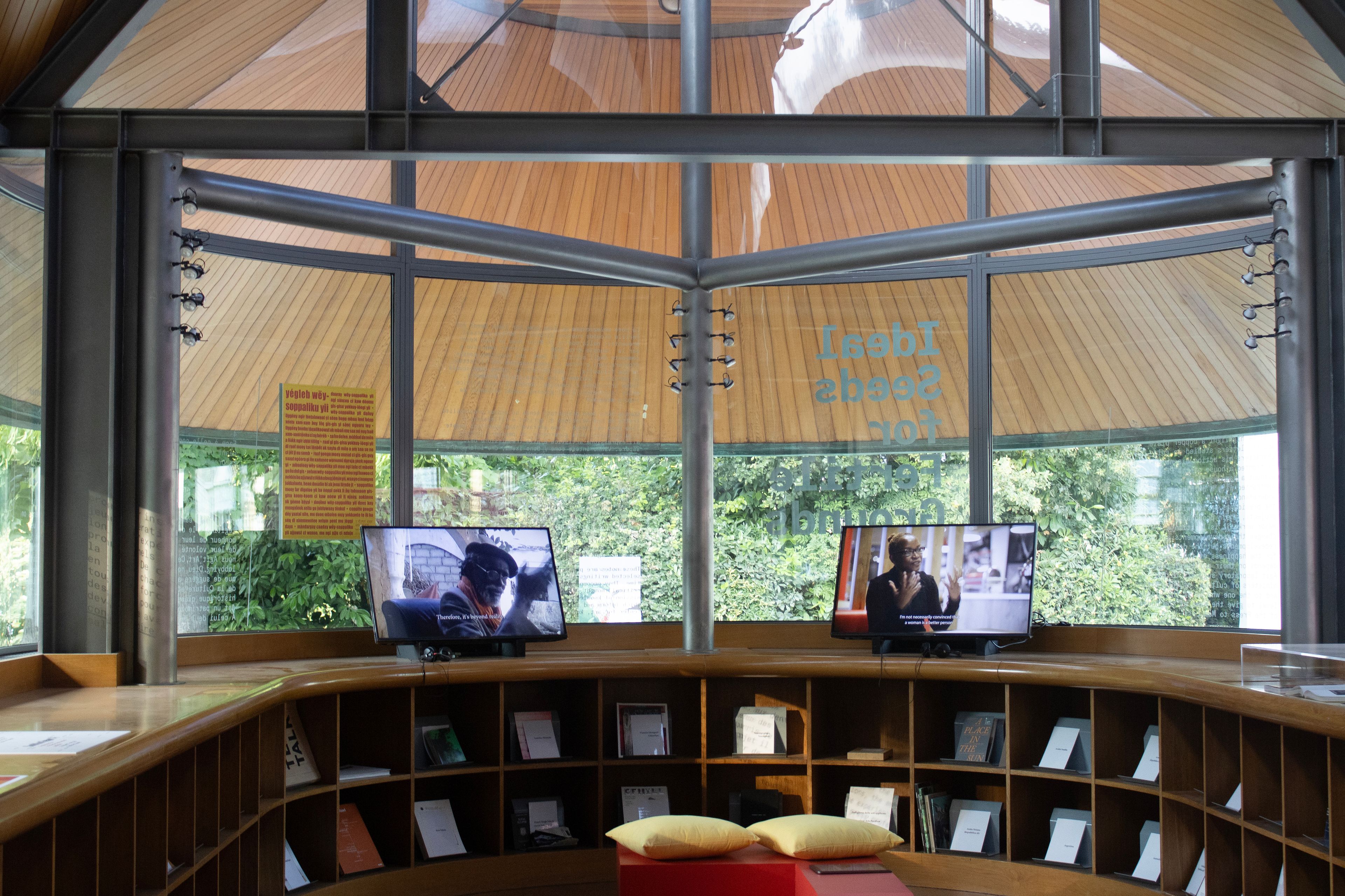 A modern, circular room with a curved wooden counter displaying books and two video screens. Large windows offer a view of trees, and the ceiling features wooden panels and exposed metal beams.