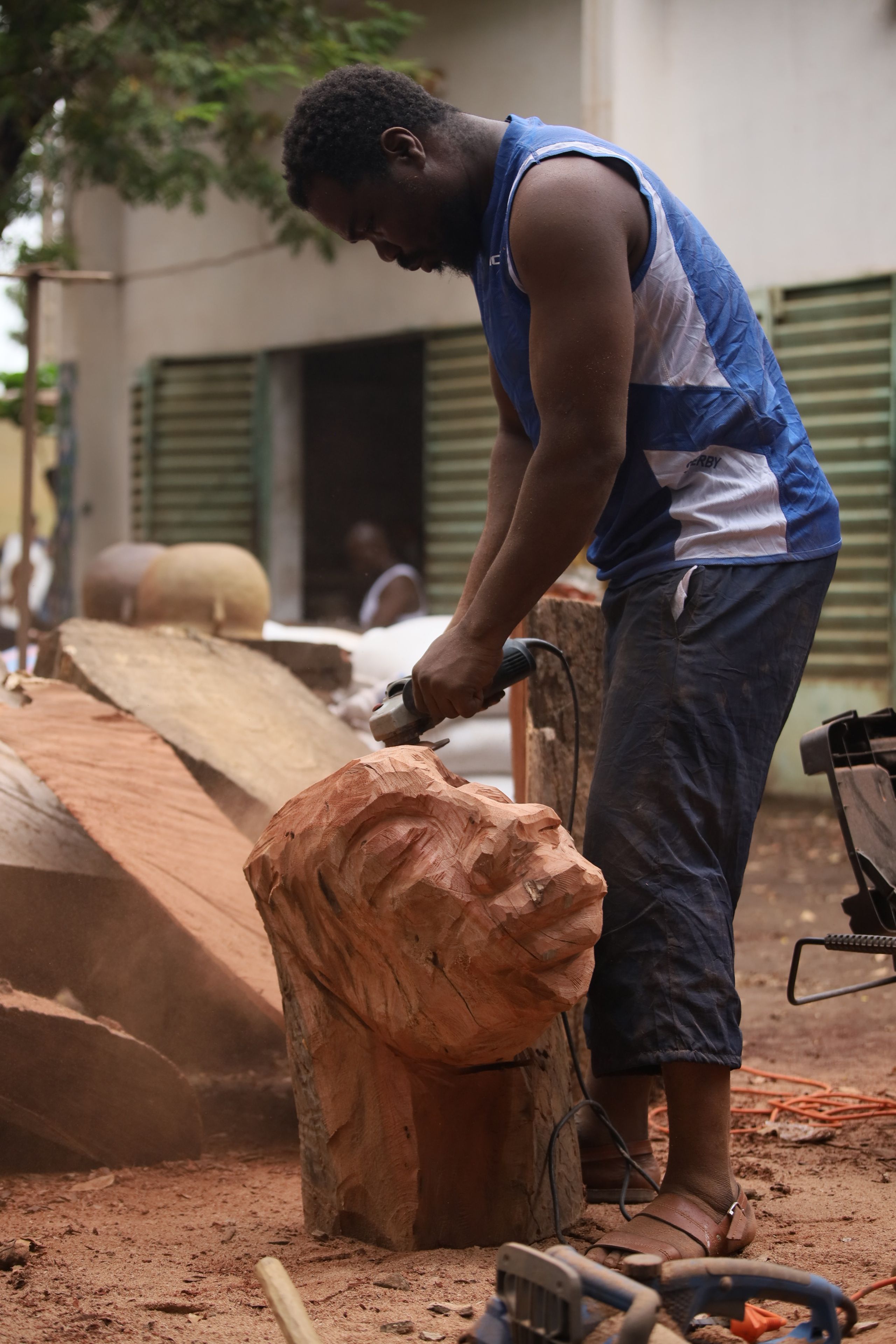 A man uses an angle grinder to carve a large wooden head sculpture outdoors.