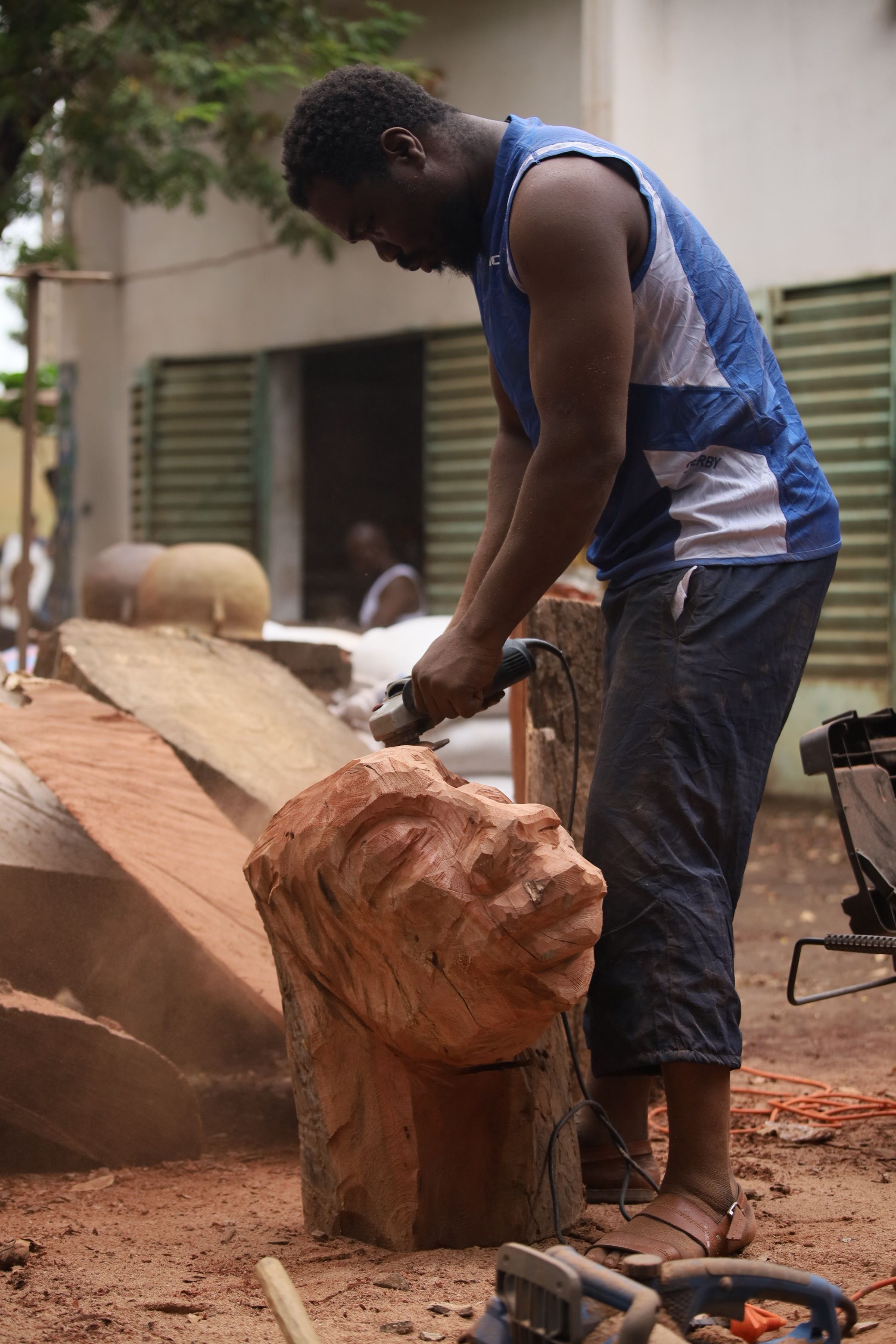 A man uses an angle grinder to carve a large wooden head sculpture outdoors.