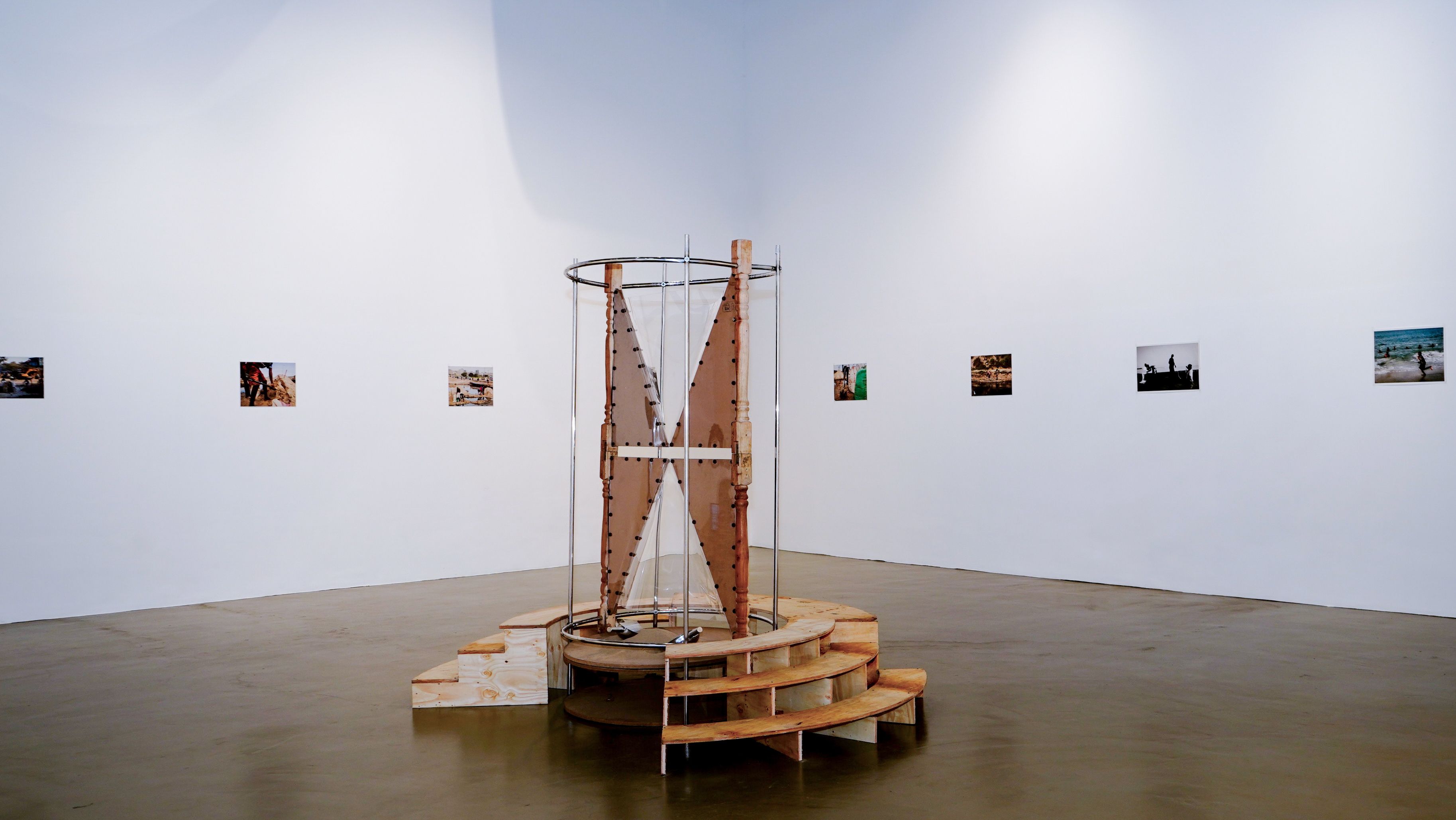 An art installation with a cylindrical wooden and metal structure on a stepped base, in front of a white wall with a row of small photographs.