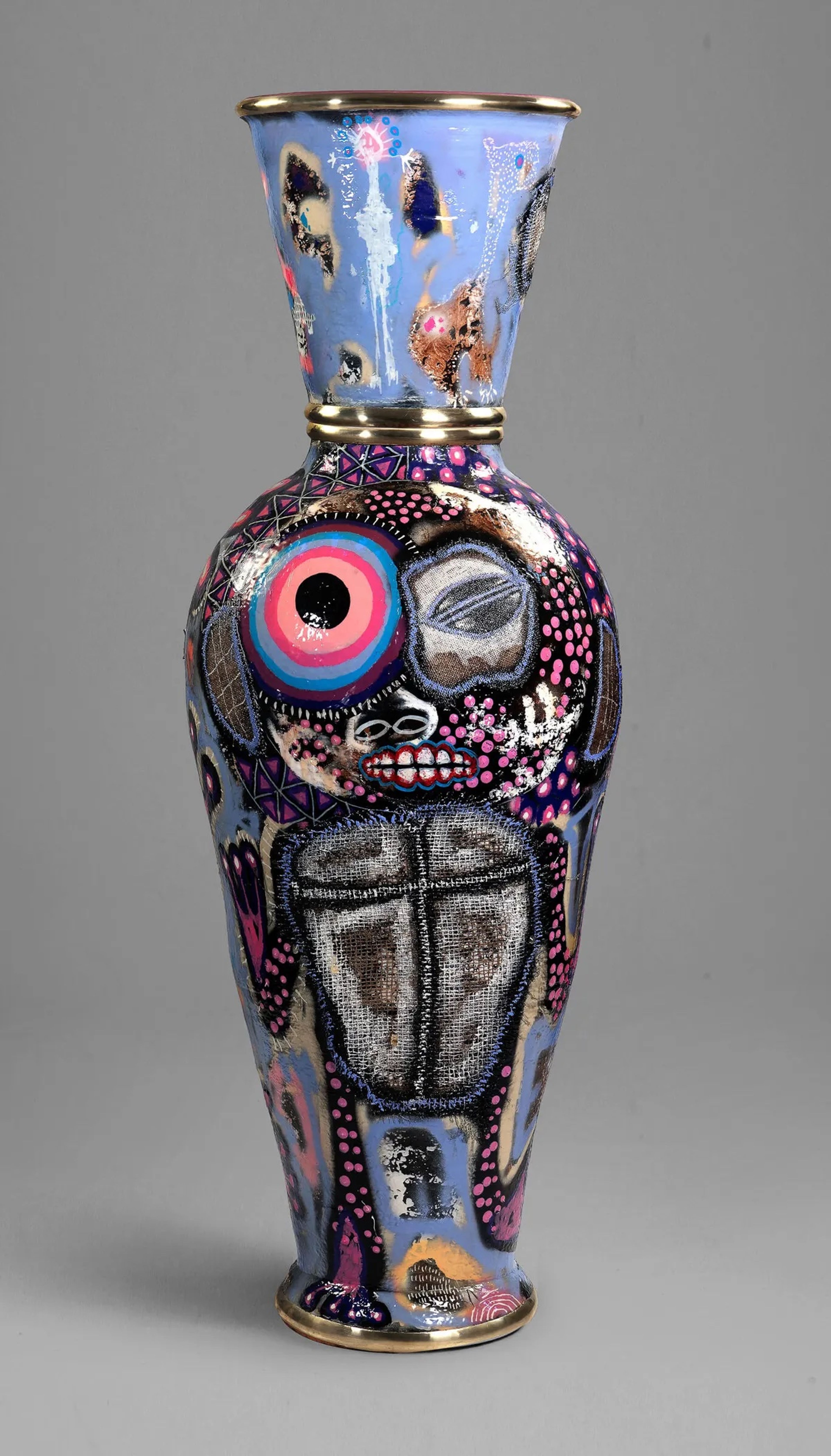 A tall, ornate vase painted with vibrant abstract patterns, including an anthropomorphic face with a large eye, mouth, and a cross on its chest, highlighted by gold bands.