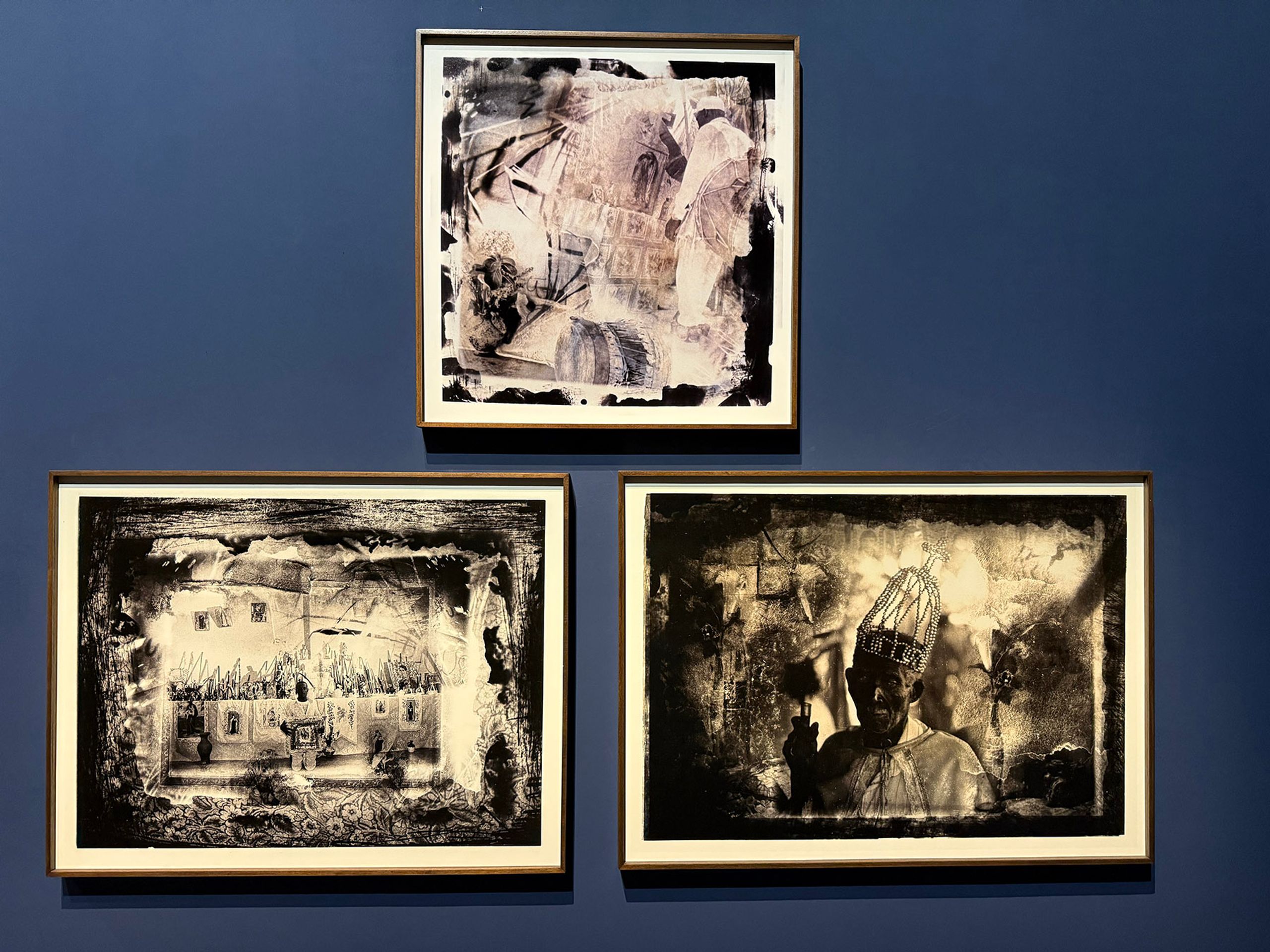 Three framed textured monochrome artworks on a dark blue wall, depicting an abstract image, a house, and a person with a birdcage hat.