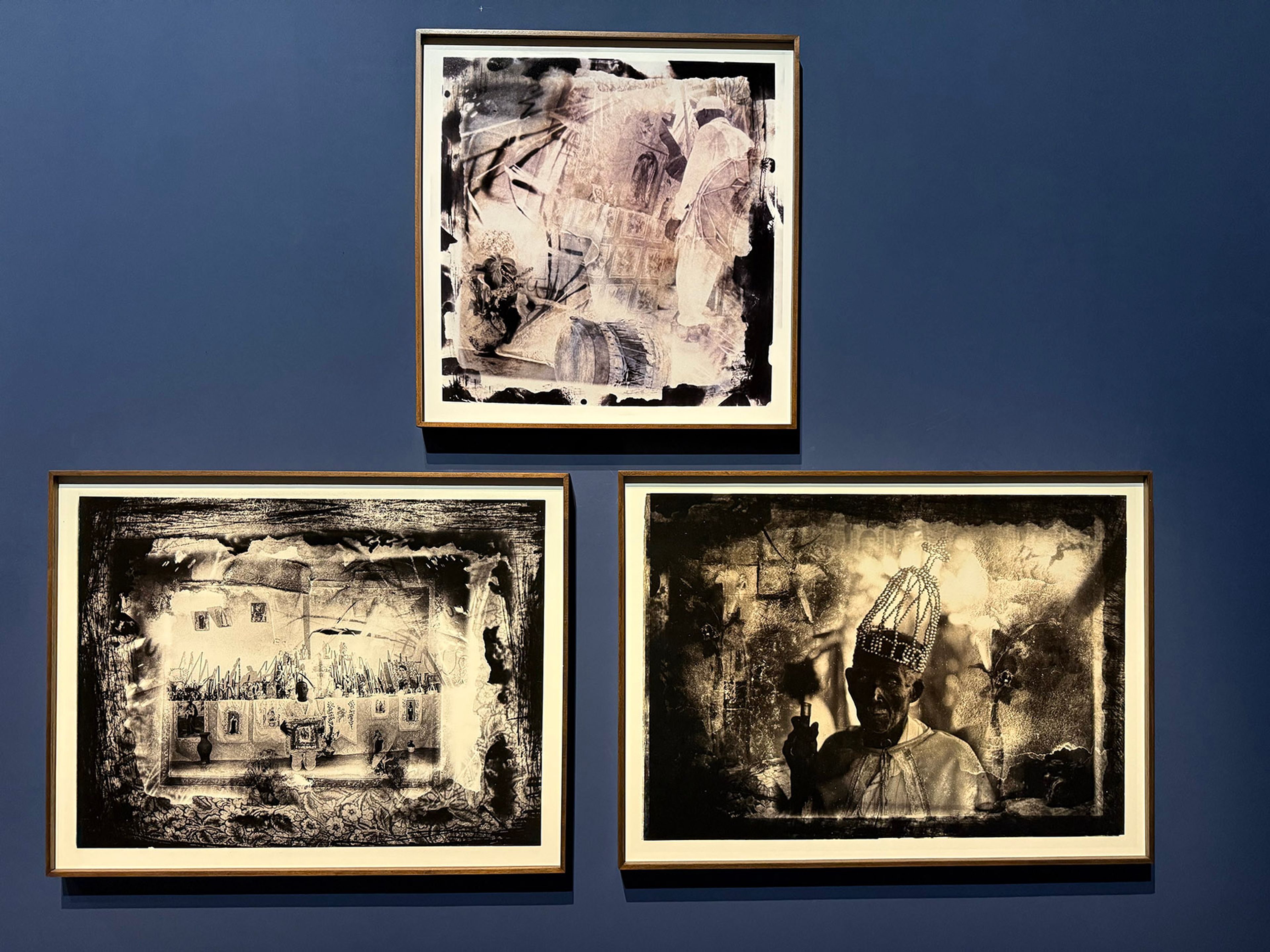 Three framed textured monochrome artworks on a dark blue wall, depicting an abstract image, a house, and a person with a birdcage hat.