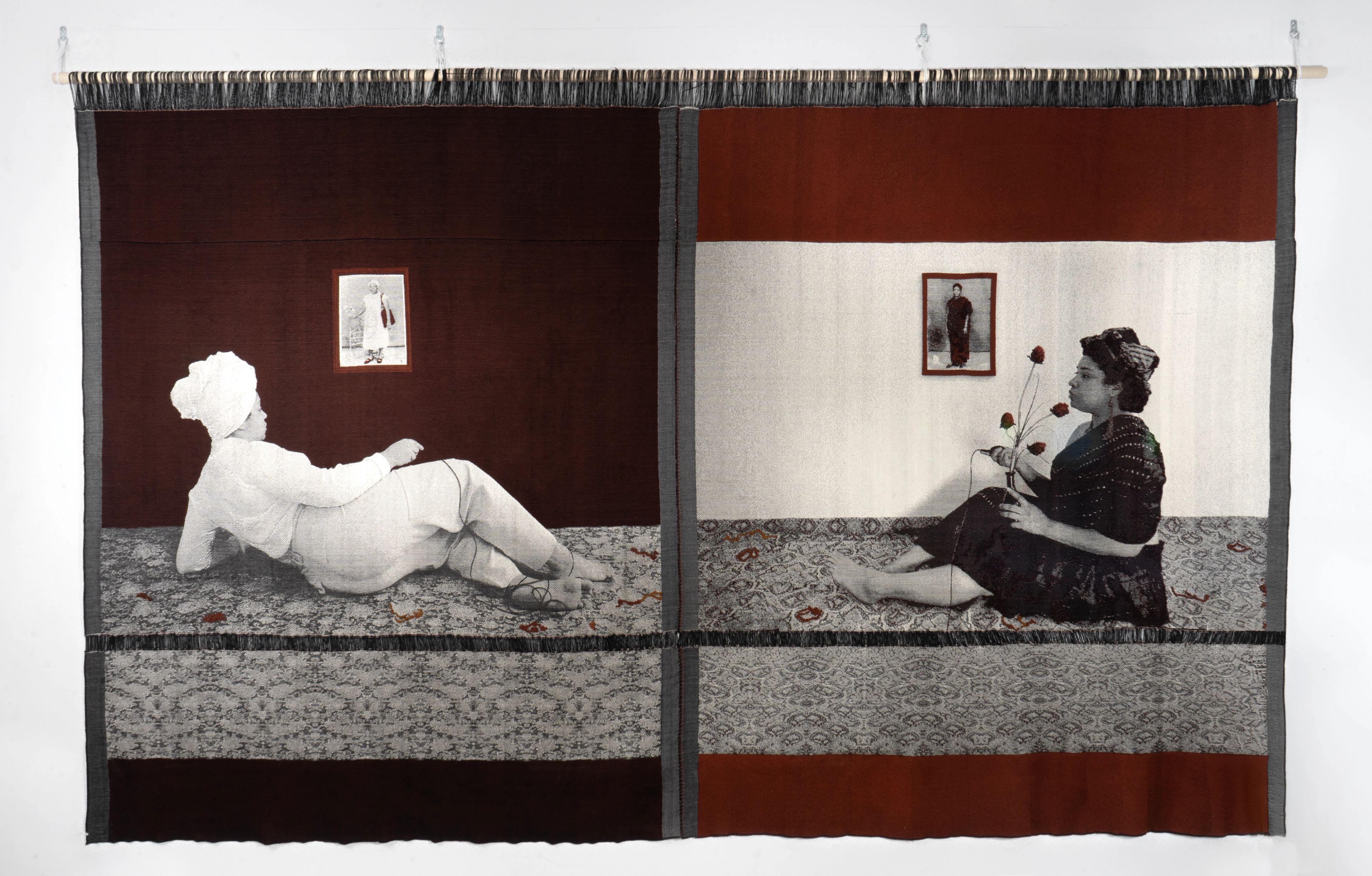 A large textile artwork showing two scenes. Left: a grayscale figure in white reclines on a patterned floor, facing a framed photo on a red wall. Right: a grayscale figure in dark clothes sits on a patterned floor, holding red flowers, facing a framed photo on a white wall.