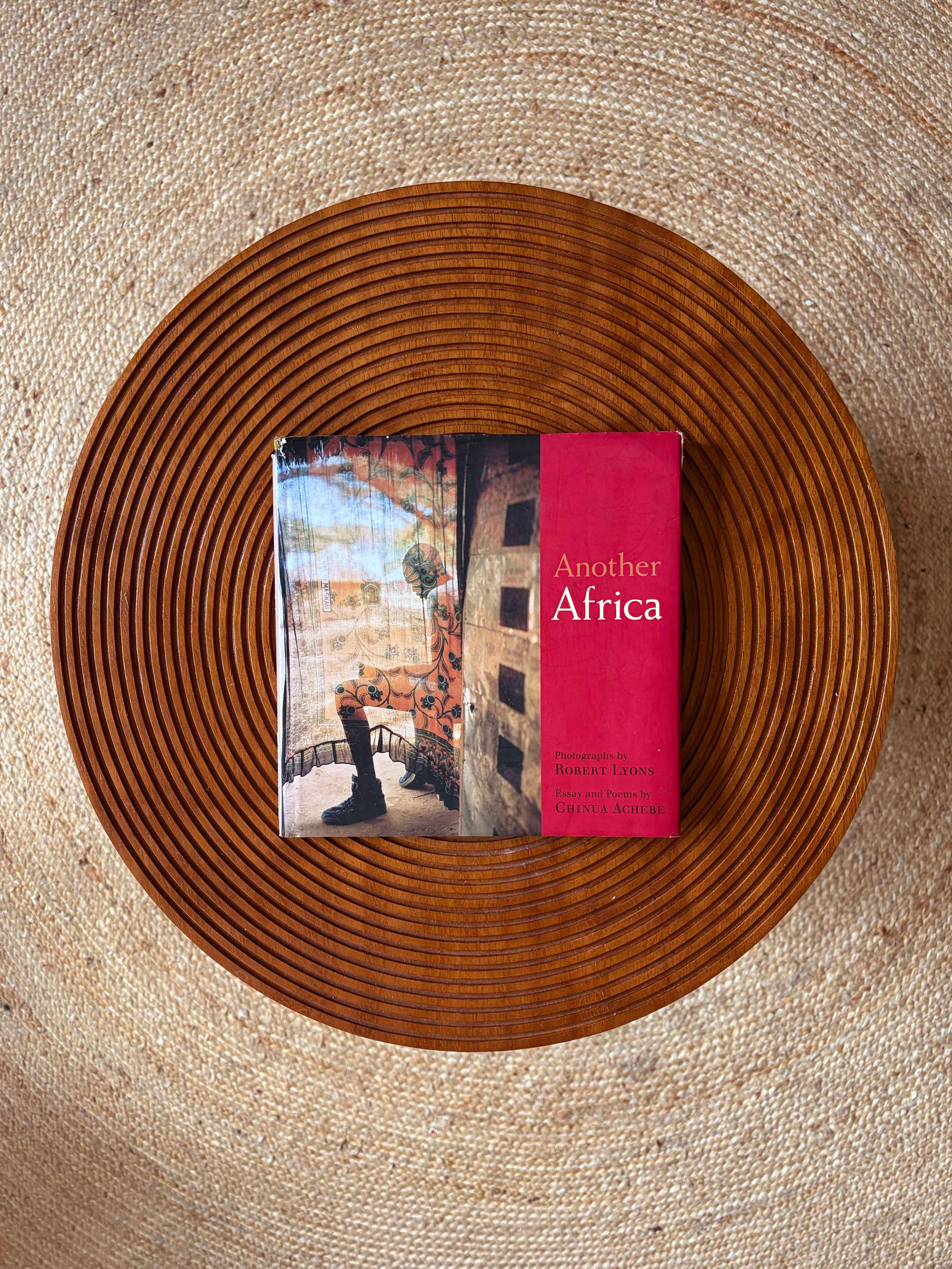 Overhead view of the book "Another Africa" on a round wooden platter atop a beige woven rug.