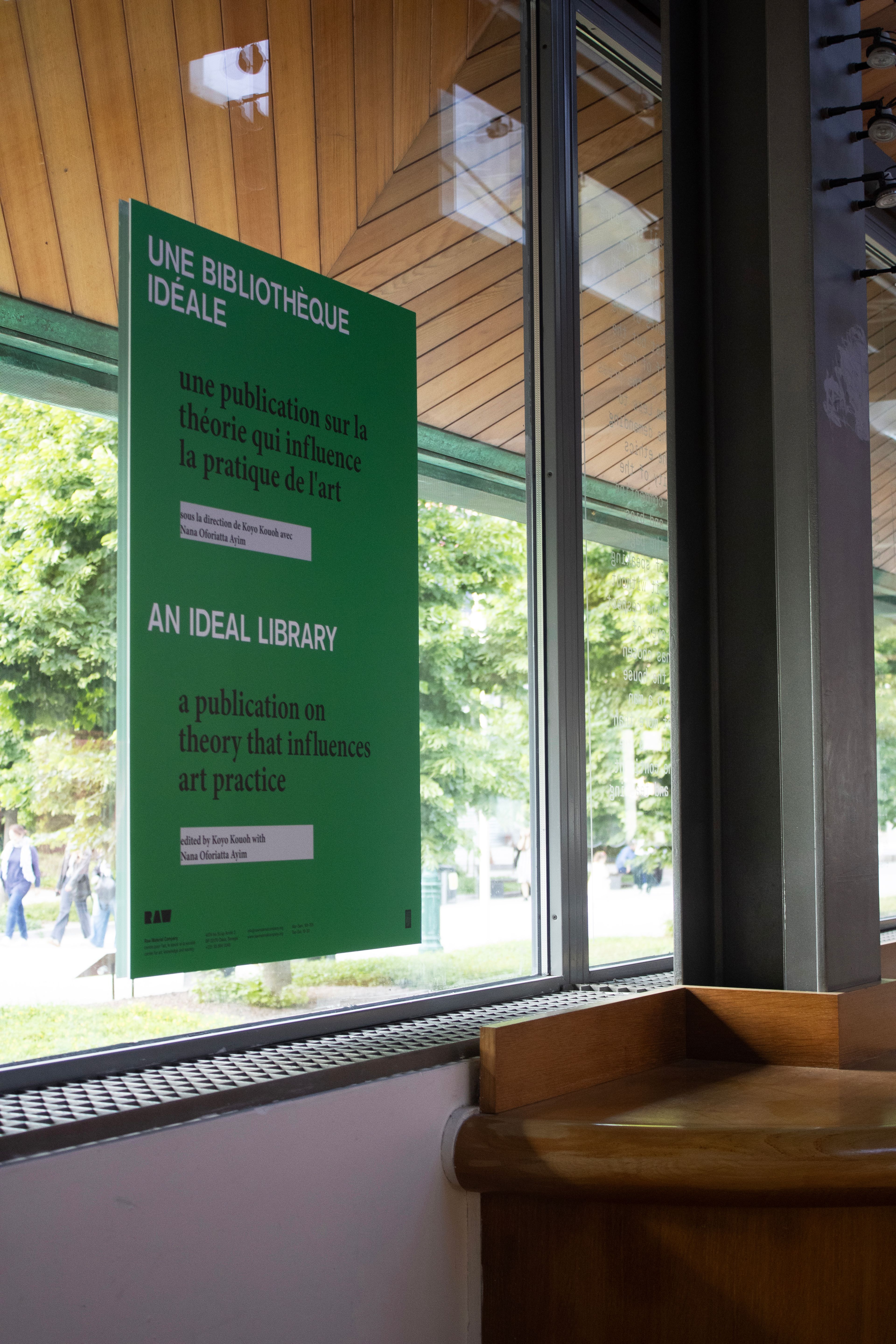 A green sign hanging in a window displays "An Ideal Library: a publication on theory that influences art practice" in English and French, with trees visible outside.
