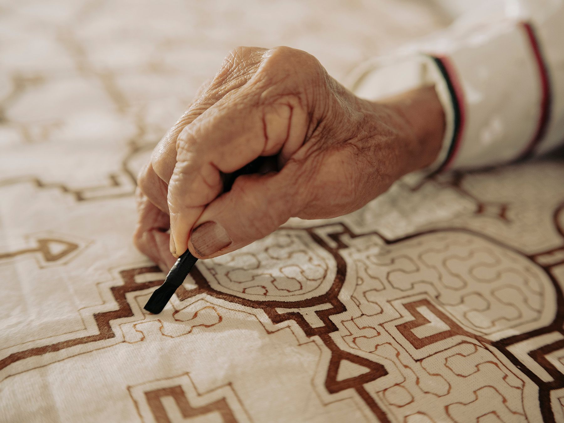 An elderly hand applies intricate brown patterns onto light fabric with a tool.