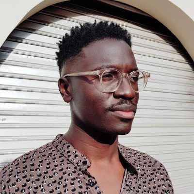 A dark-skinned man with short textured hair, a mustache, and clear glasses, wearing a patterned shirt, looks right against a corrugated metal background.