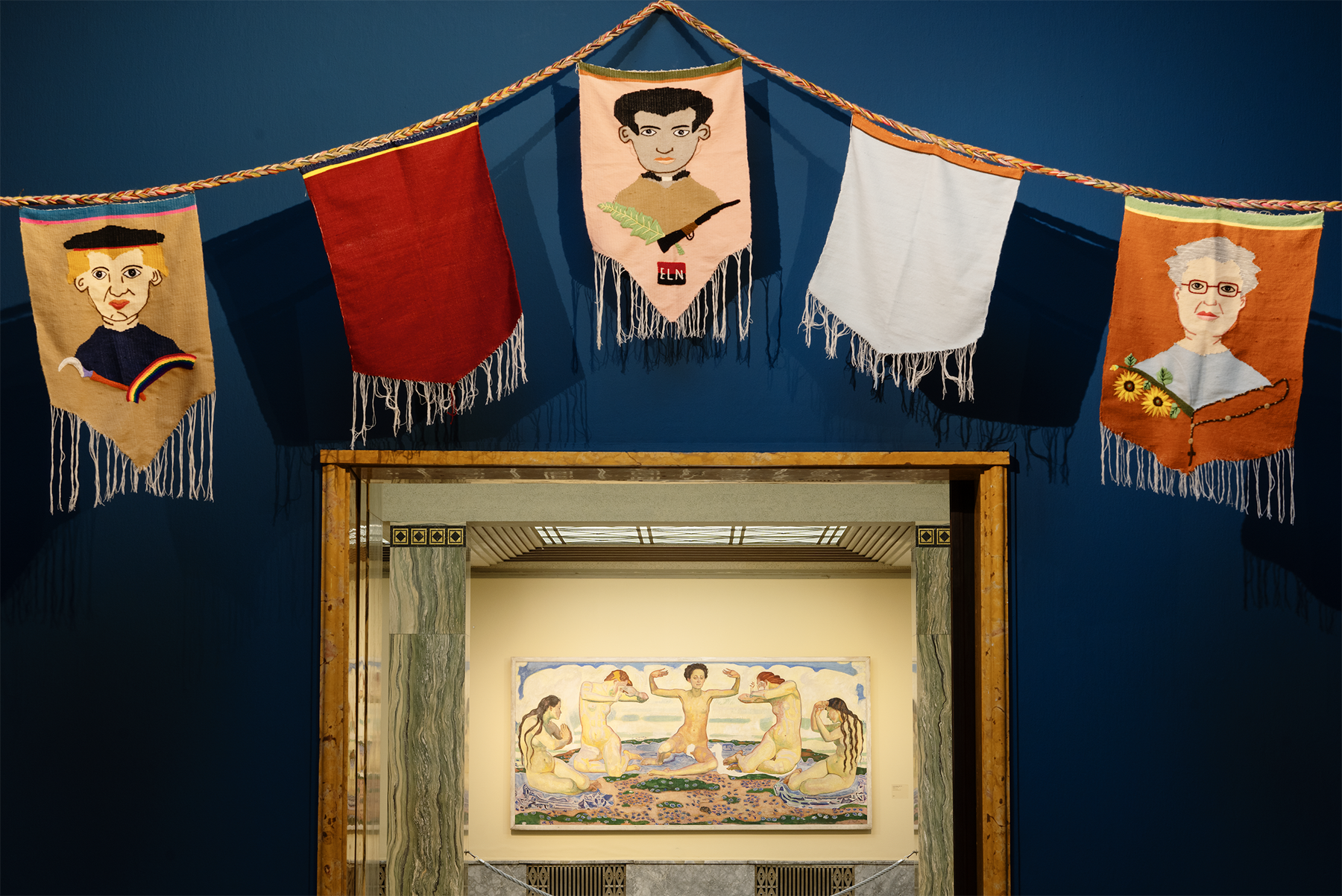 A garland of five textile banners, some with embroidered portraits and others plain, hangs on a blue wall above an open doorway revealing a painting in the room beyond.