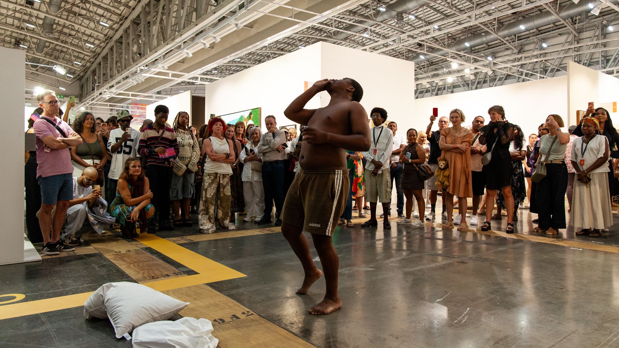A shirtless Black man performs barefoot, drinking from a small object, while a diverse crowd watches in an art exhibition hall.