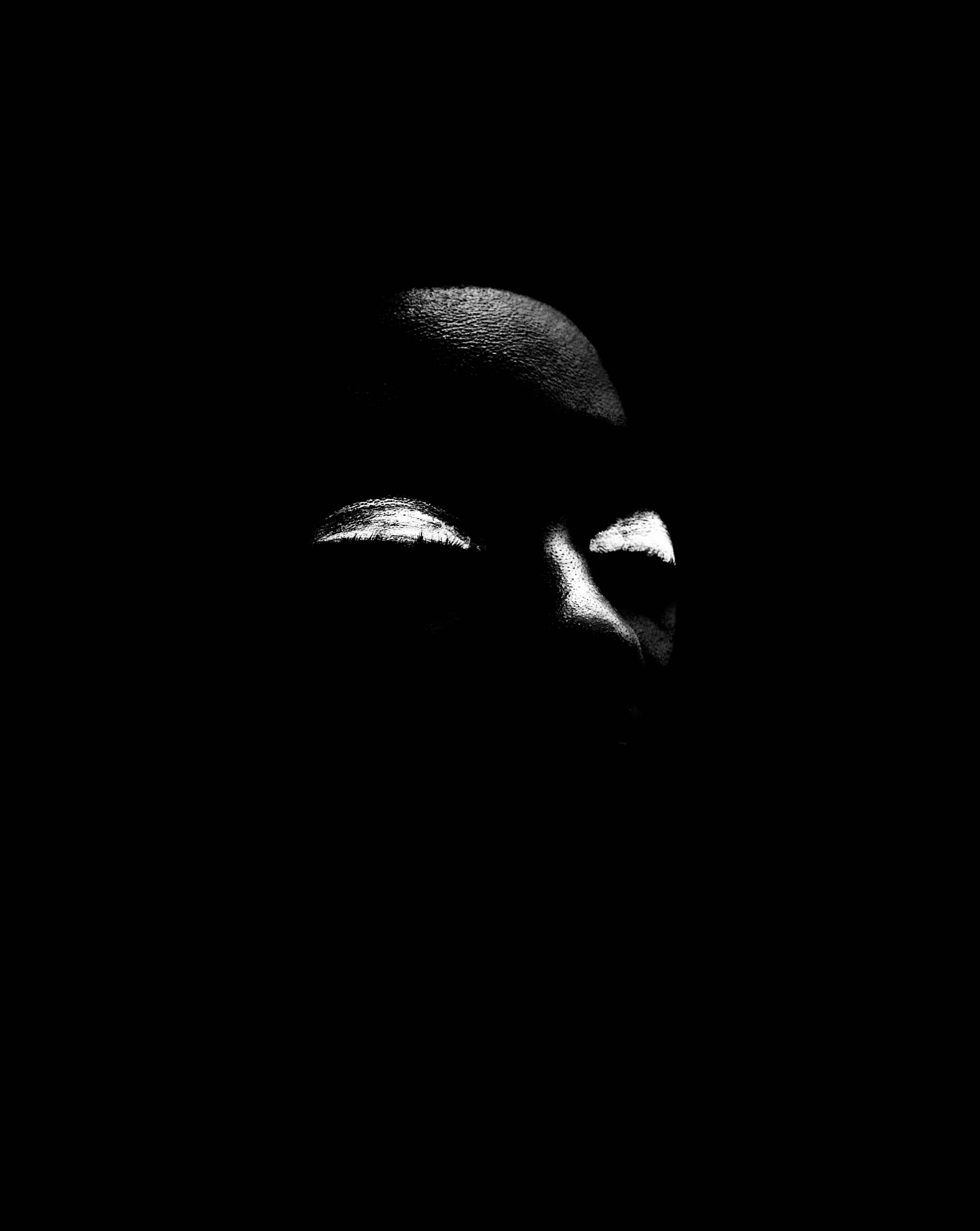 Stark black and white close-up of a face, with the forehead, closed eyelids, and nose ridge brightly highlighted against a dark background.