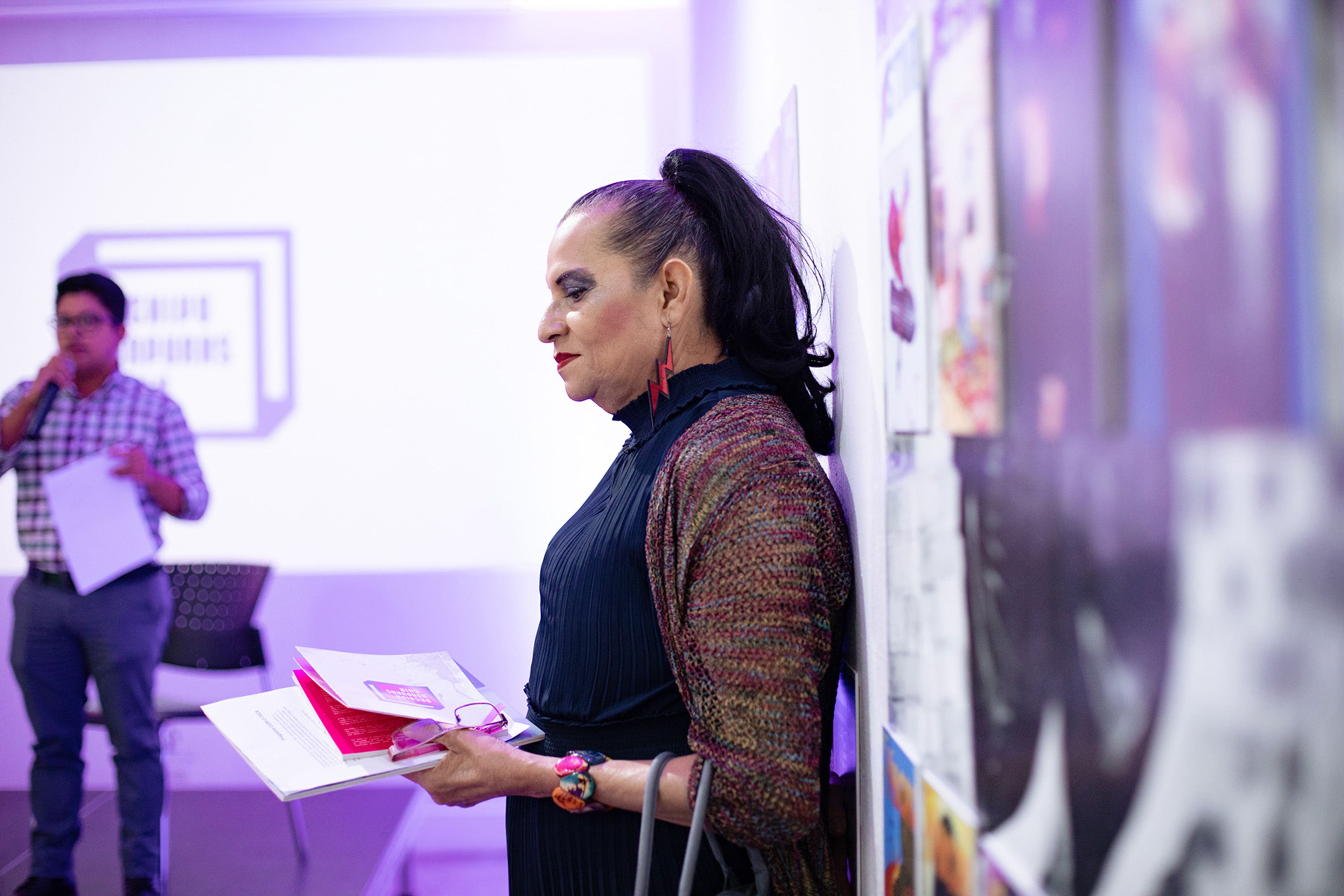 A woman with dark hair pulled back, wearing a colorful cardigan and red earrings, looks down at a book she holds. A man with a microphone is in the background.