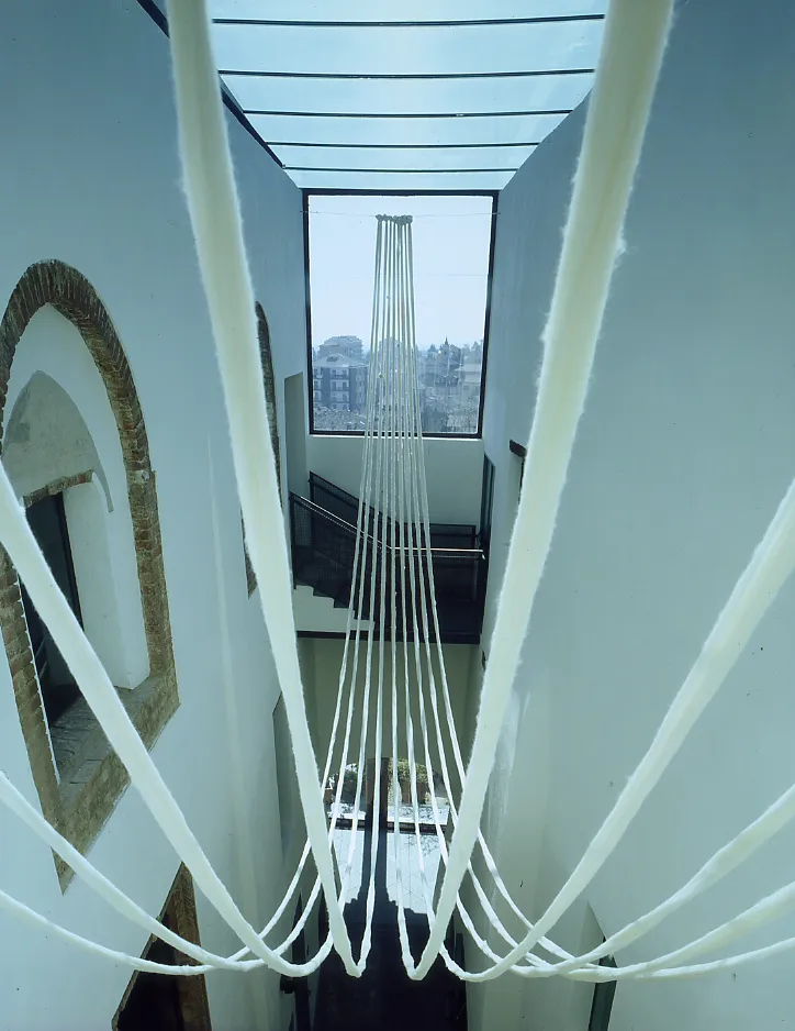 An art installation of white fabric ropes hangs within a bright atrium with a glass ceiling, some looping in the foreground, others vertical against a large window overlooking a city.