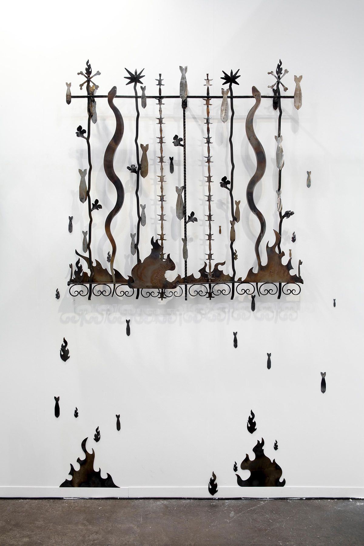 Metal wall sculpture depicting a fence of snakes, barbed wire, and bombs, with additional bomb and flame shapes falling onto a white wall.