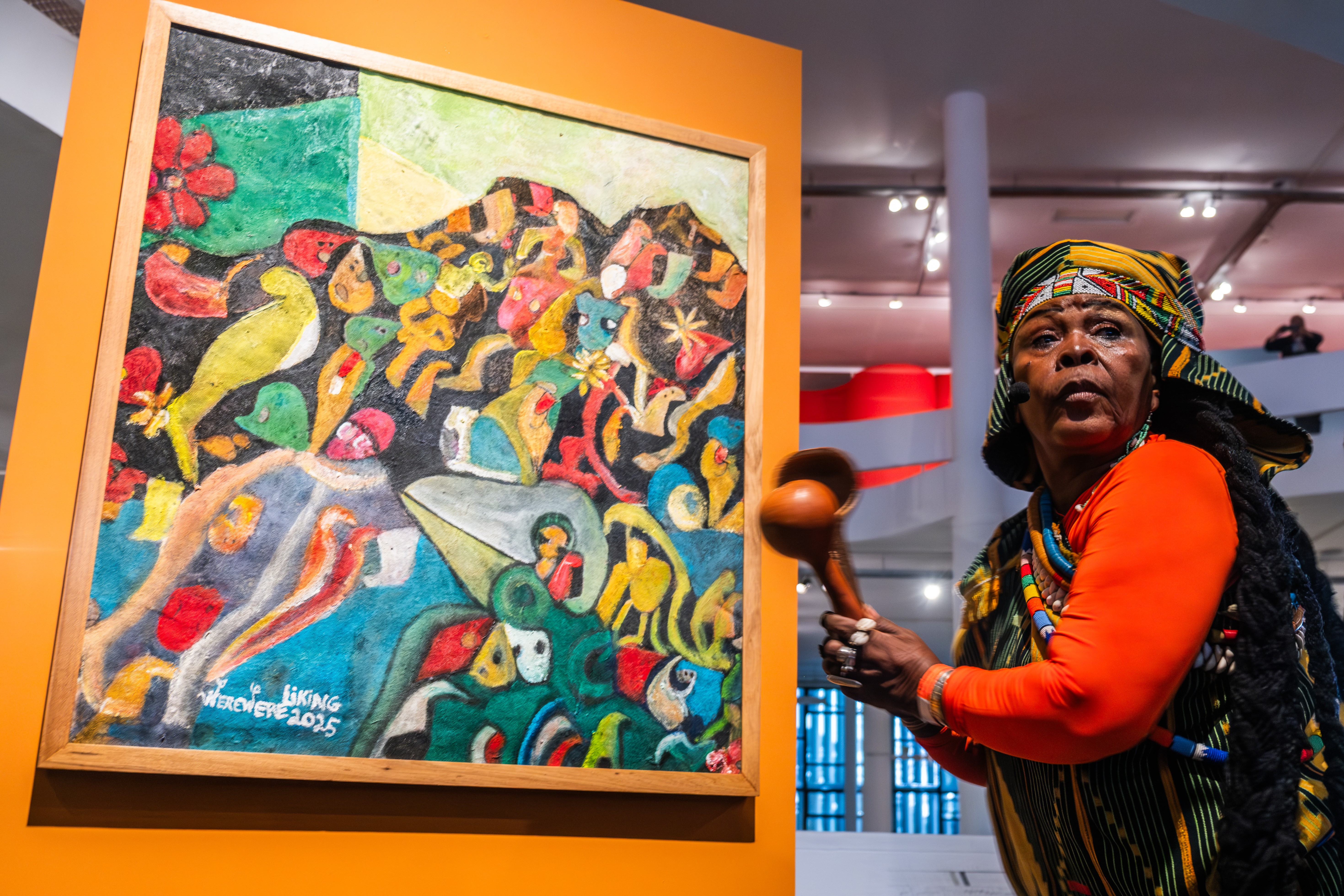 A woman in a colorful headwrap holds a gourd rattle next to a vibrant abstract painting.
