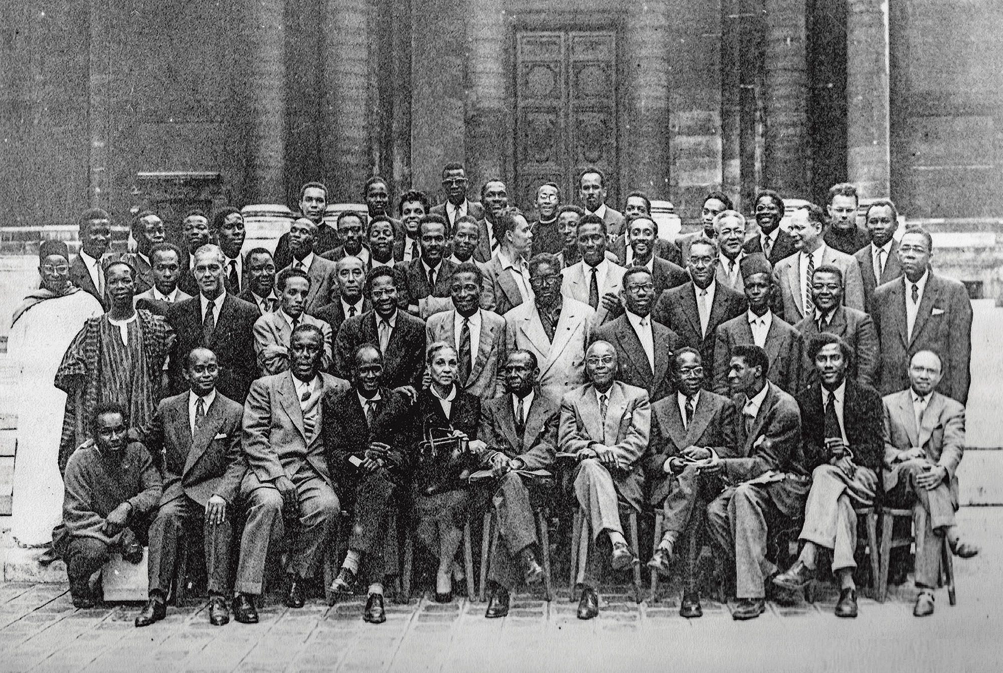 Formal black and white group portrait of dozens of men, mostly Black, and one woman, in suits and traditional clothing, gathered outside a building.
