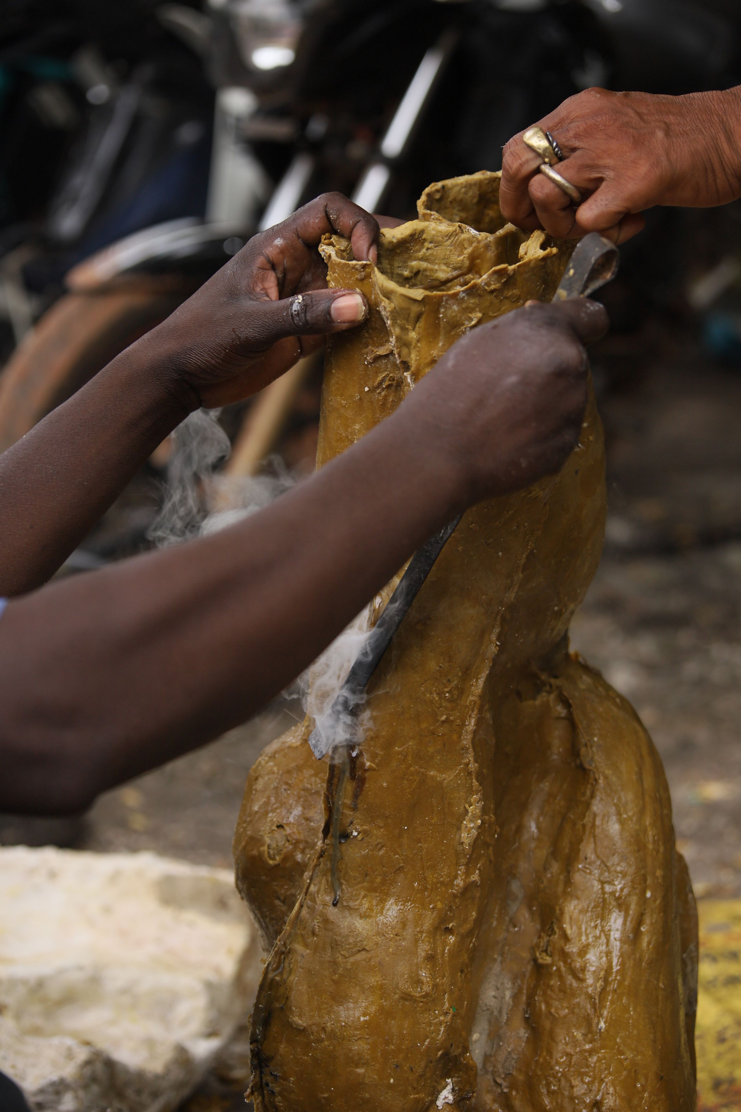 Hands shaping a large, golden-brown wax-like form, with smoke rising from a tool cutting into it.