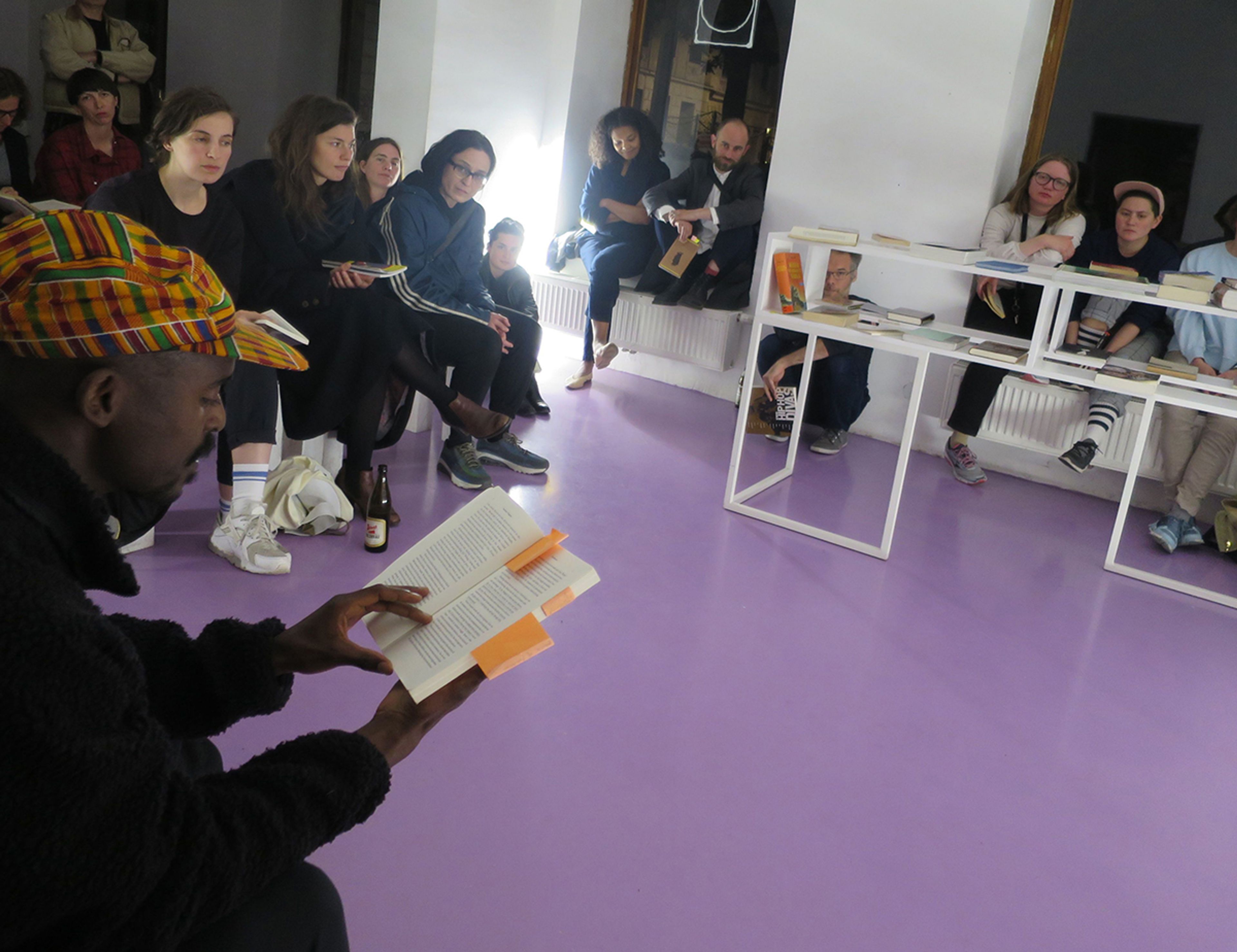 A man in a colorful plaid hat reads a tabbed book to a seated audience in a room with a purple floor and a white bookshelf.