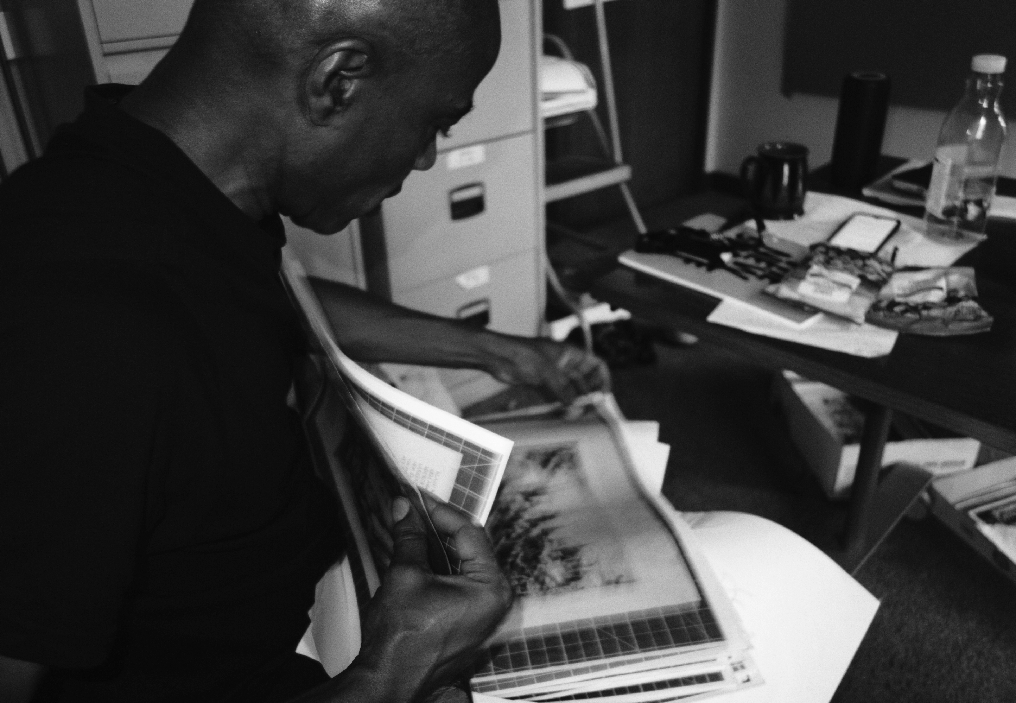 Black and white photo of a man looking through prints in a cluttered office.