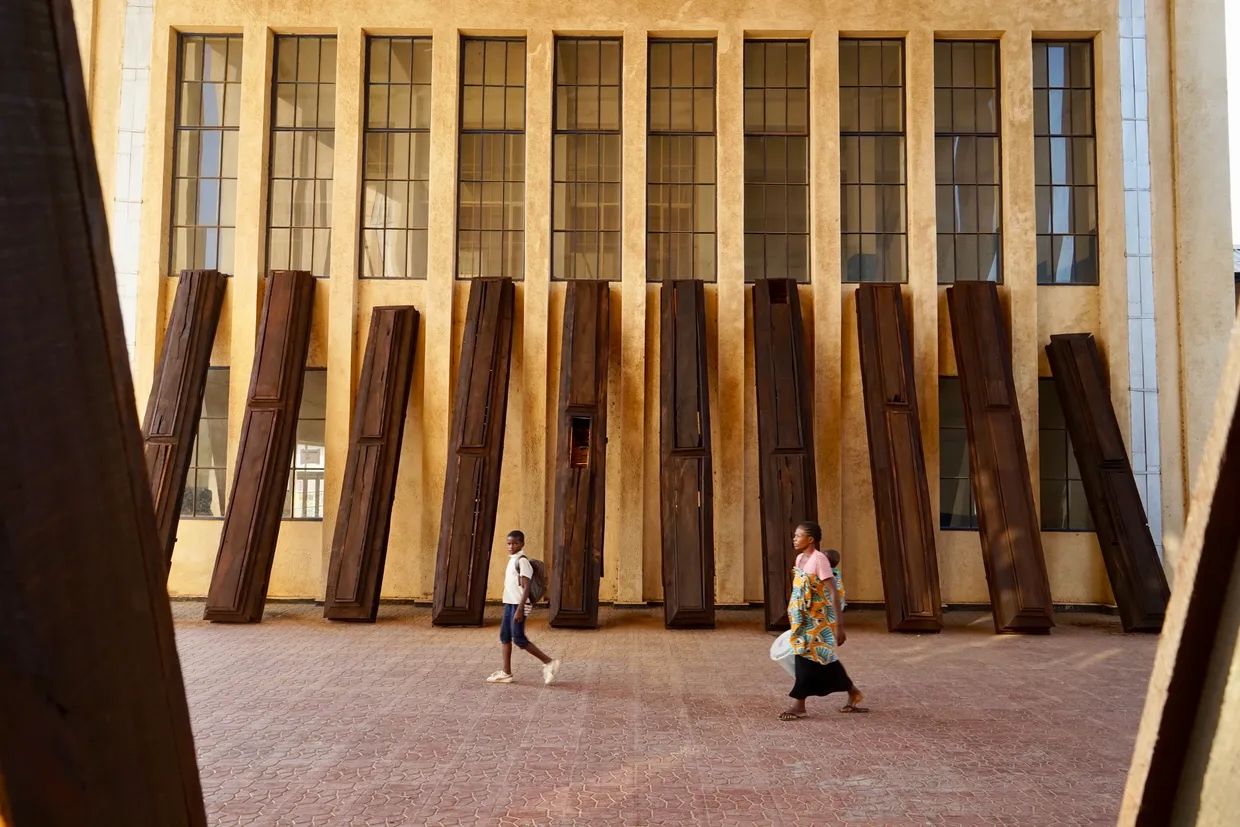 Two people walk past a beige building with tall windows and a row of dark, coffin-like structures leaning against its wall.
