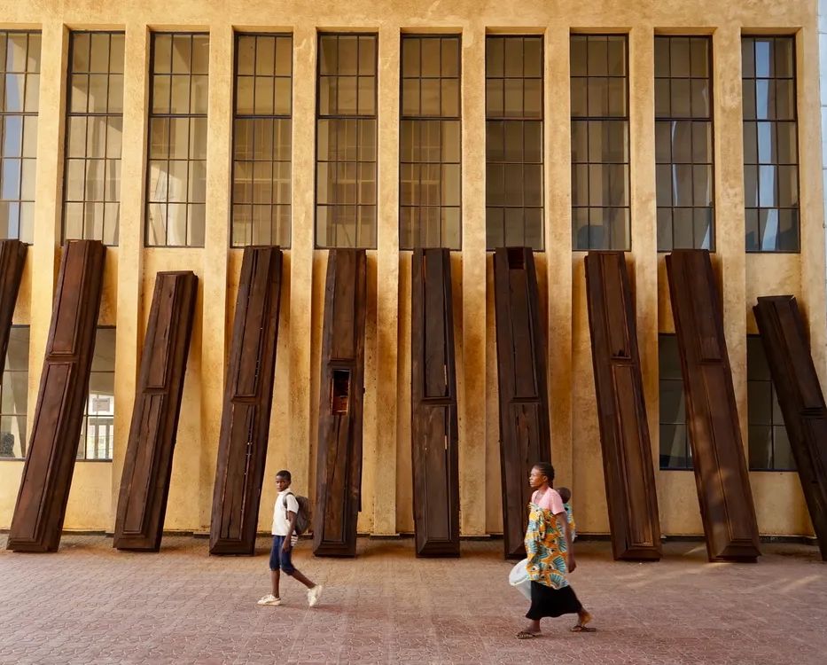 Two people walk past a beige building with tall windows and a row of dark, coffin-like structures leaning against its wall.