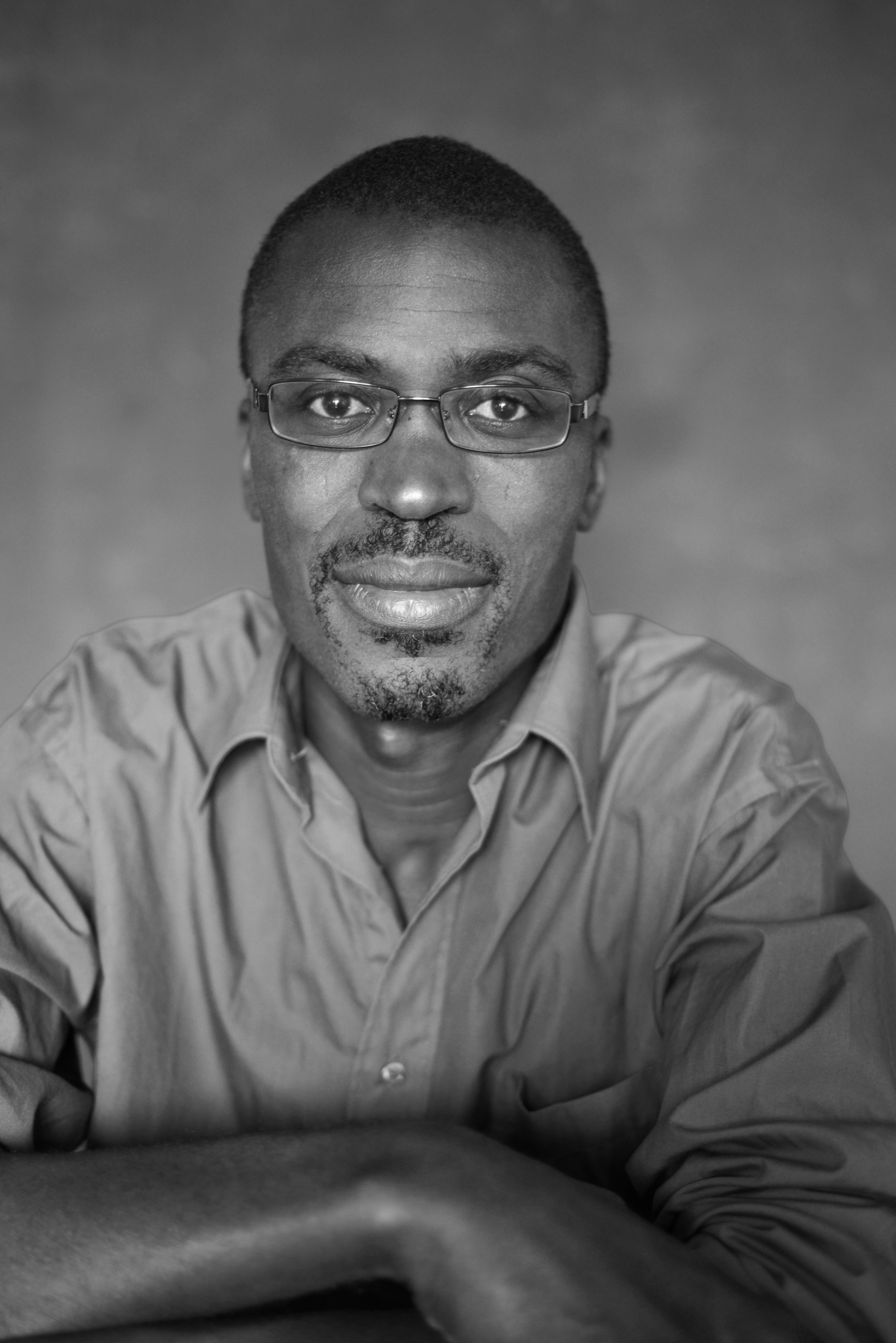 Black and white mid-shot of a Black man with glasses and a collared shirt, looking at the viewer.