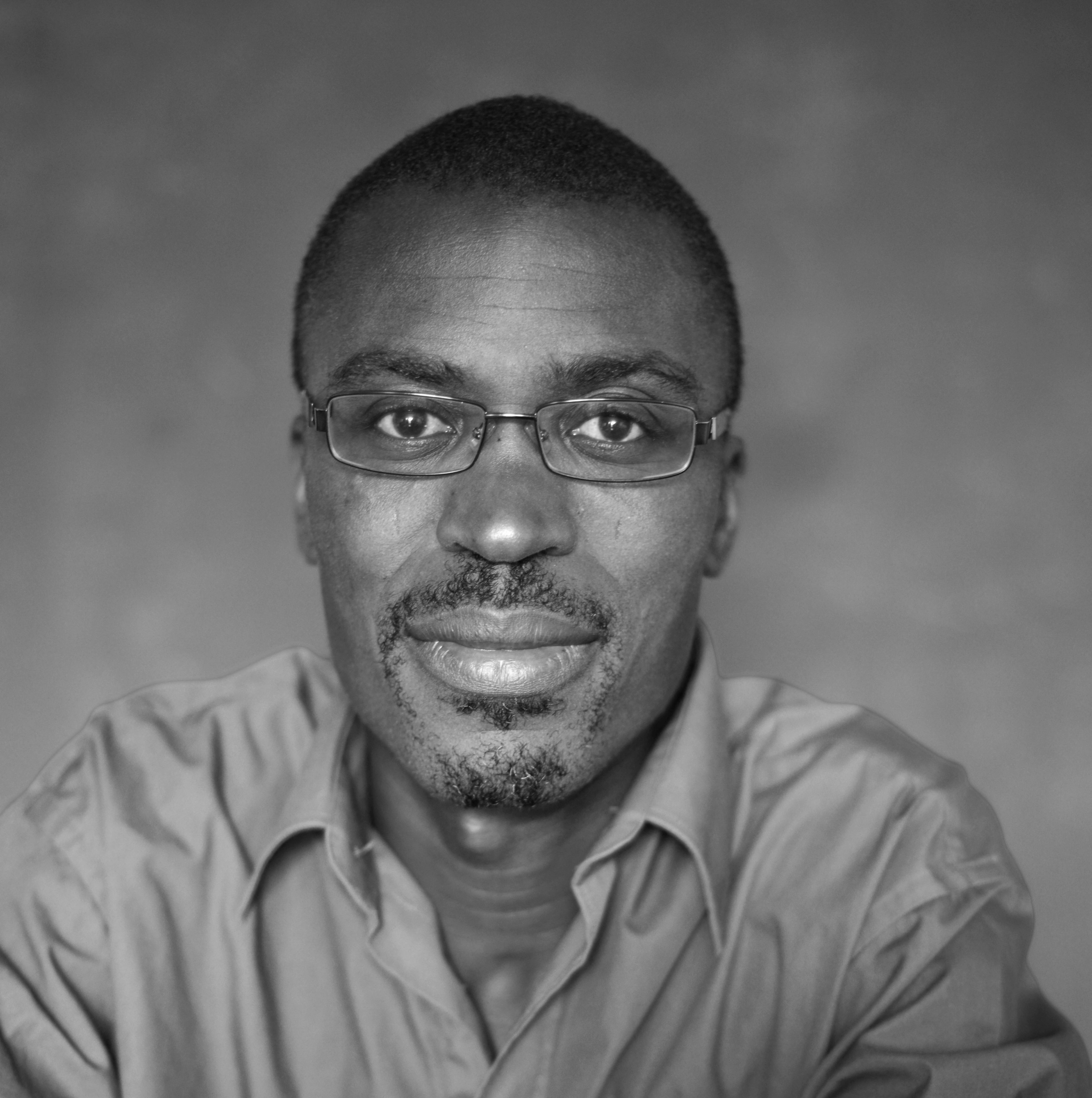 Black and white mid-shot of a Black man with glasses and a collared shirt, looking at the viewer.