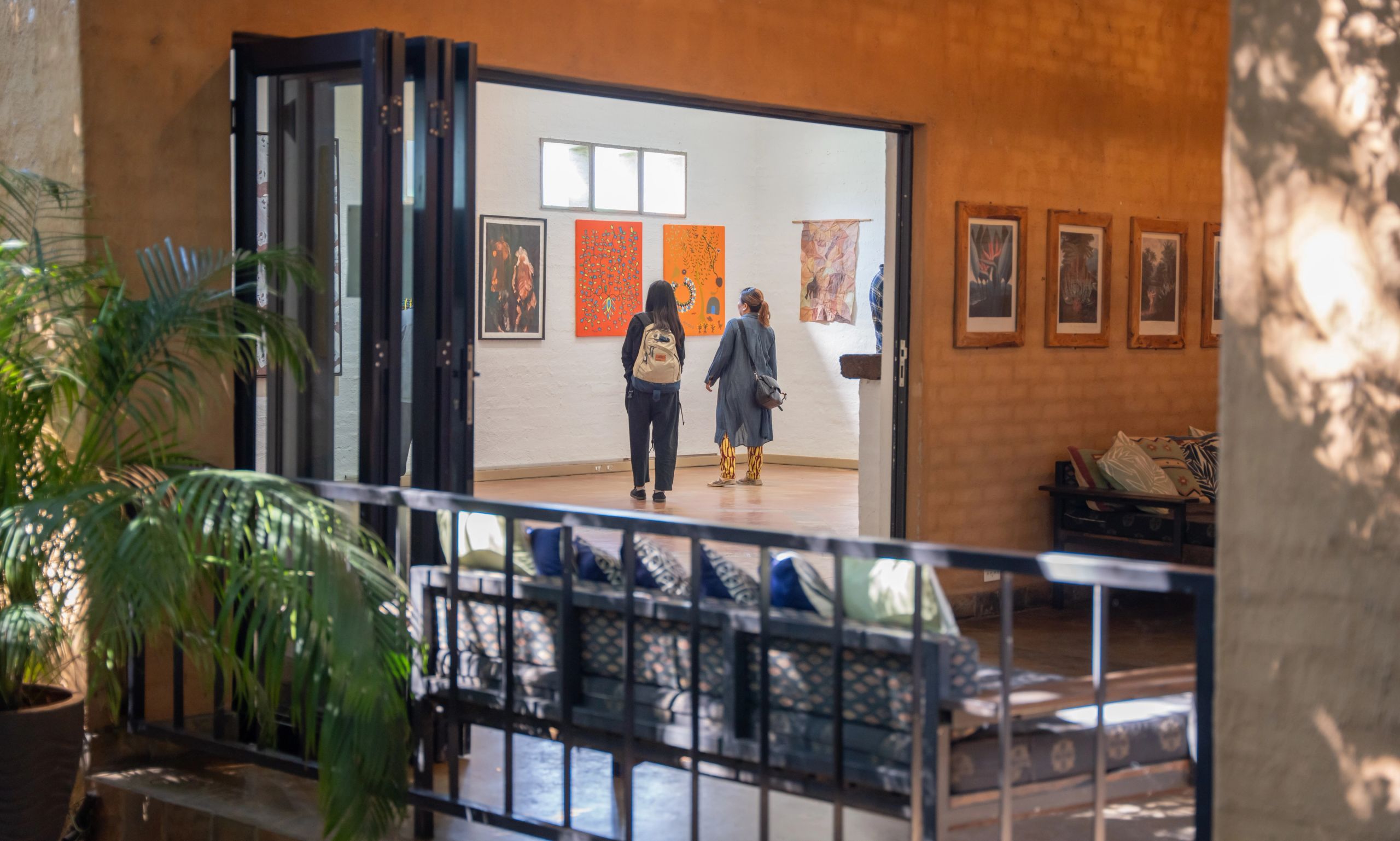 Two people look at vibrant paintings in a bright art gallery, framed by a lounge with plants and terracotta walls.