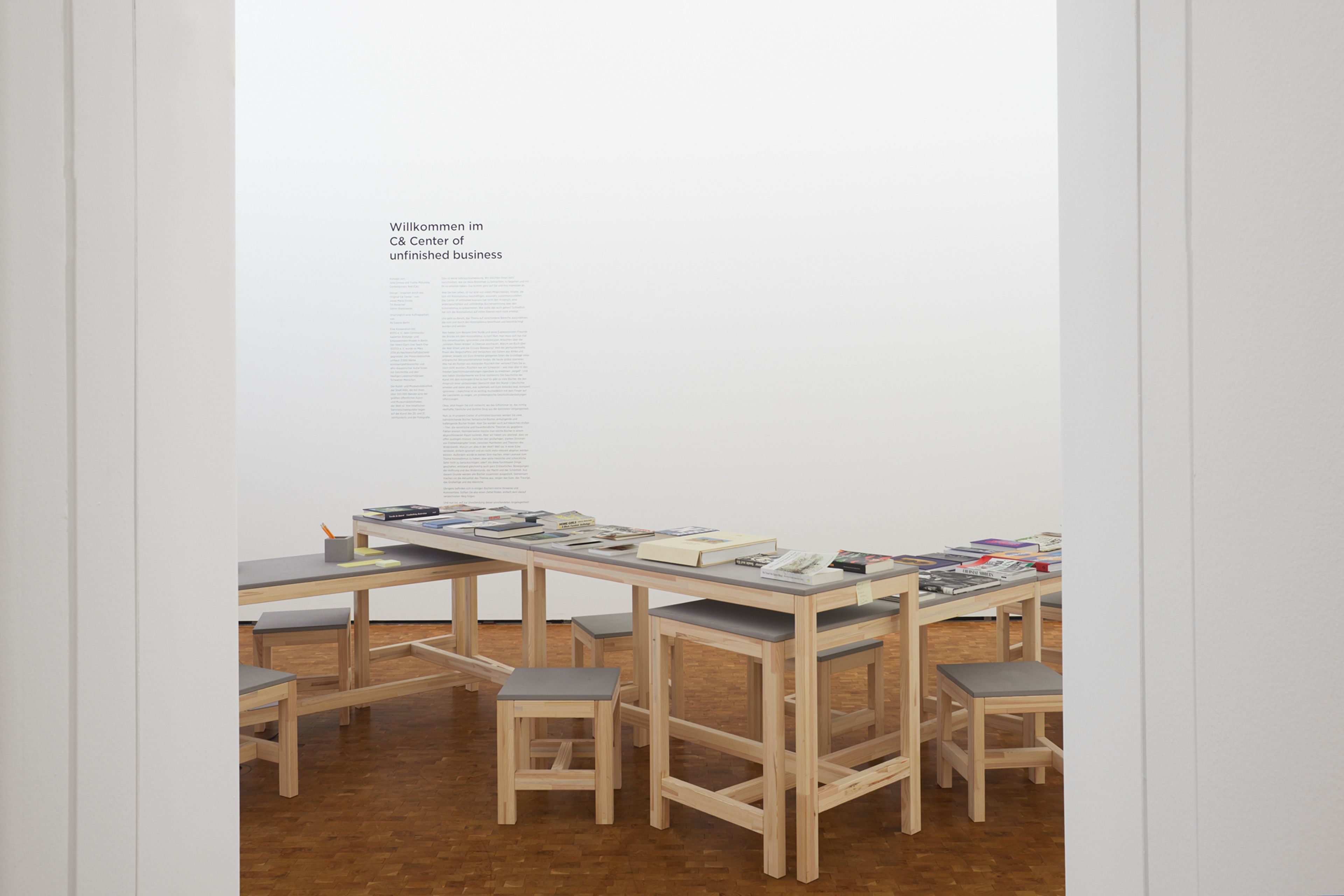 A minimalist room features a long light wood table covered with books and papers, surrounded by stools with grey tops, and white wall text reading "Willkommen im CB Center of unfinished business."