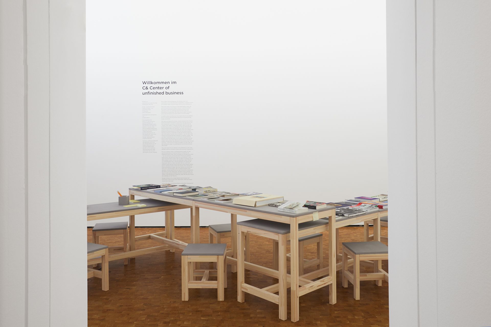 A minimalist room features a long light wood table covered with books and papers, surrounded by stools with grey tops, and white wall text reading "Willkommen im CB Center of unfinished business."