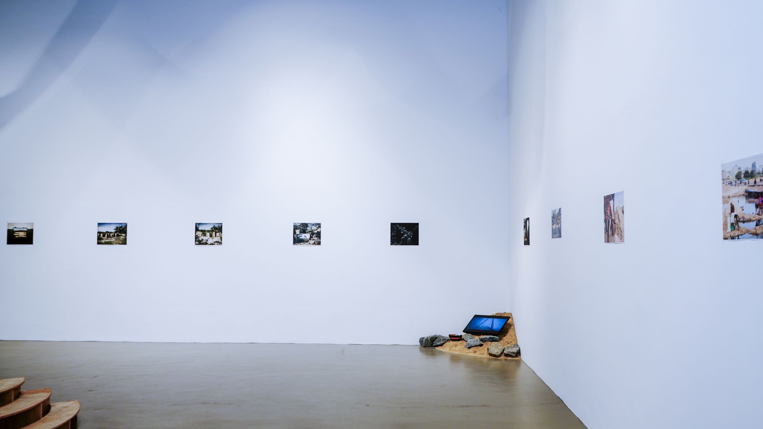 A bright art gallery featuring white walls, several framed photographs, and a floor installation with sand, rocks, and a digital screen.
