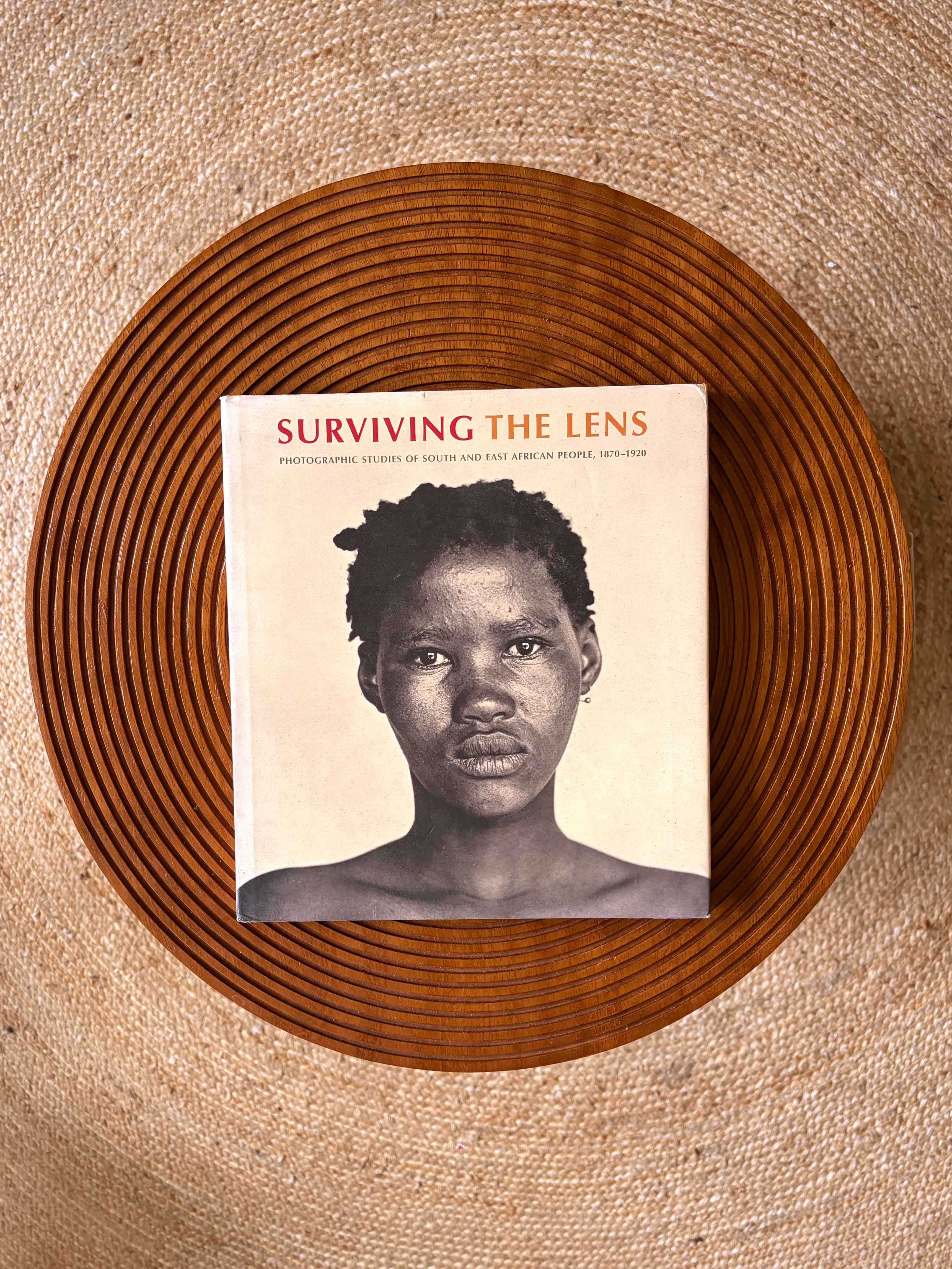 Book titled 'Surviving the Lens' with a black and white portrait of a woman on the cover, on a wooden tray on a woven mat.