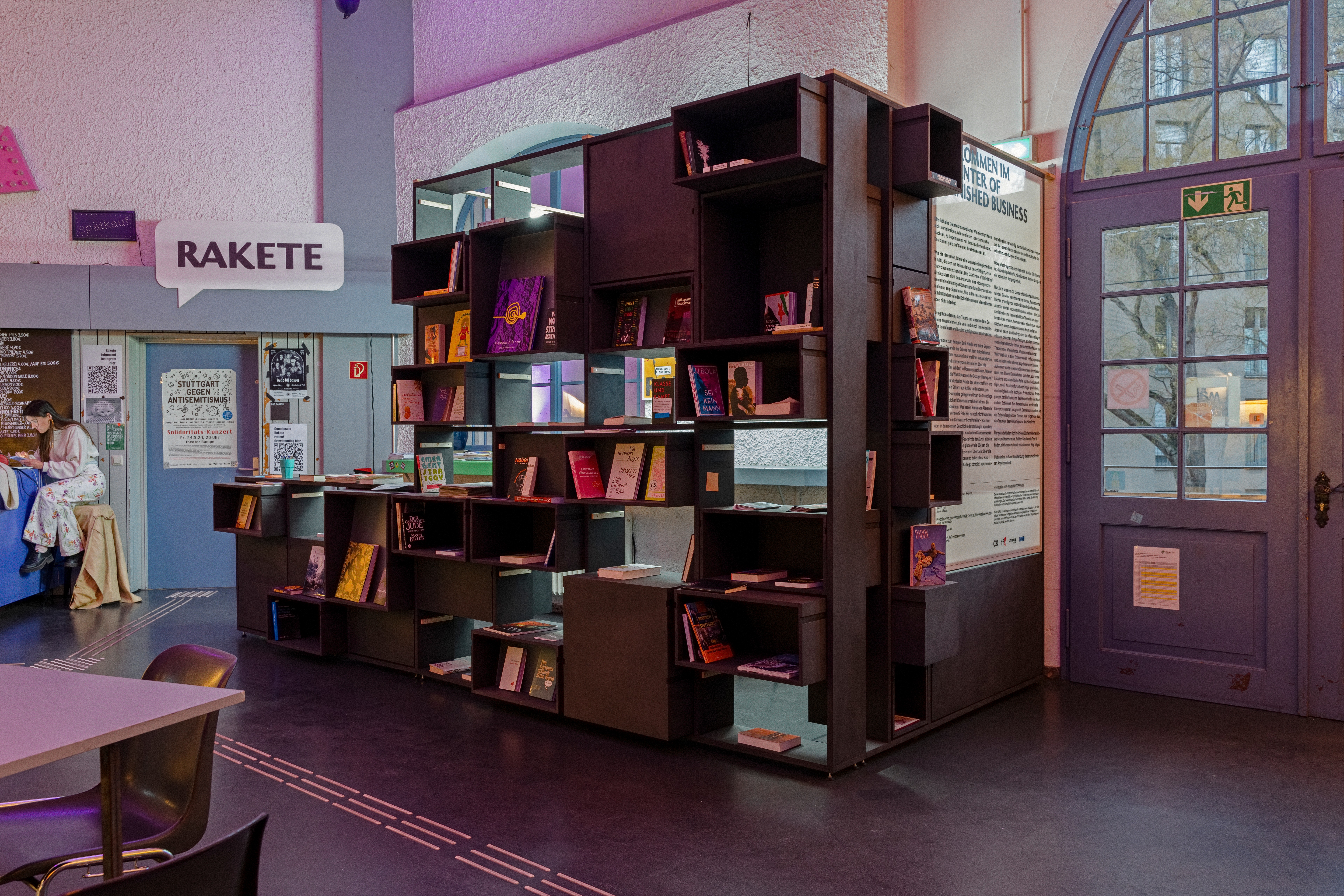 A large, dark modular bookshelf filled with books in a room with a "RAKETE" sign and an arched glass door.