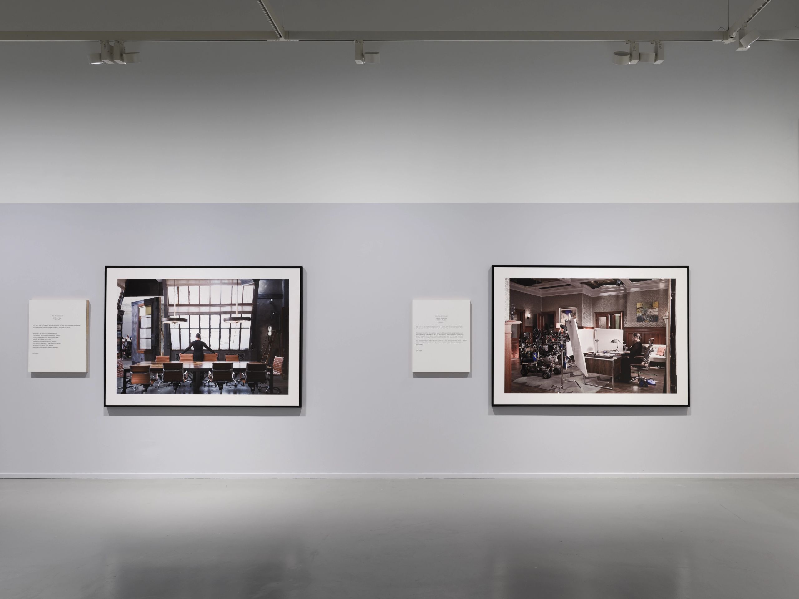 An exhibition view showing two large framed photographs on a light grey wall, one of a boardroom, the other of an interior scene with equipment.