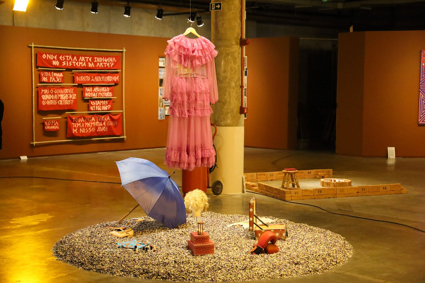An art exhibition featuring a hanging pink ruffled dress, a circular display of objects including a blue umbrella, and red banners with text on a terracotta wall.