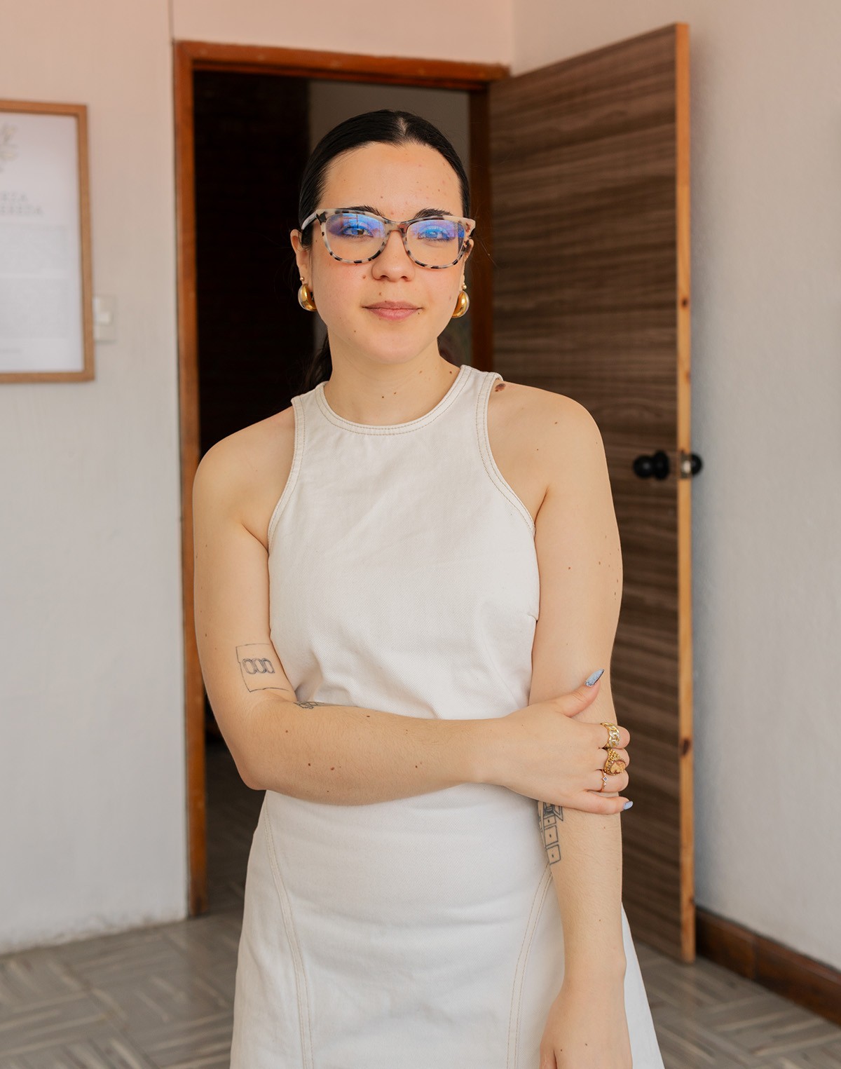 A woman with dark hair, patterned glasses, and gold hoop earrings wears a white sleeveless dress and tattoos on her arms, standing in a doorway and looking at the camera.