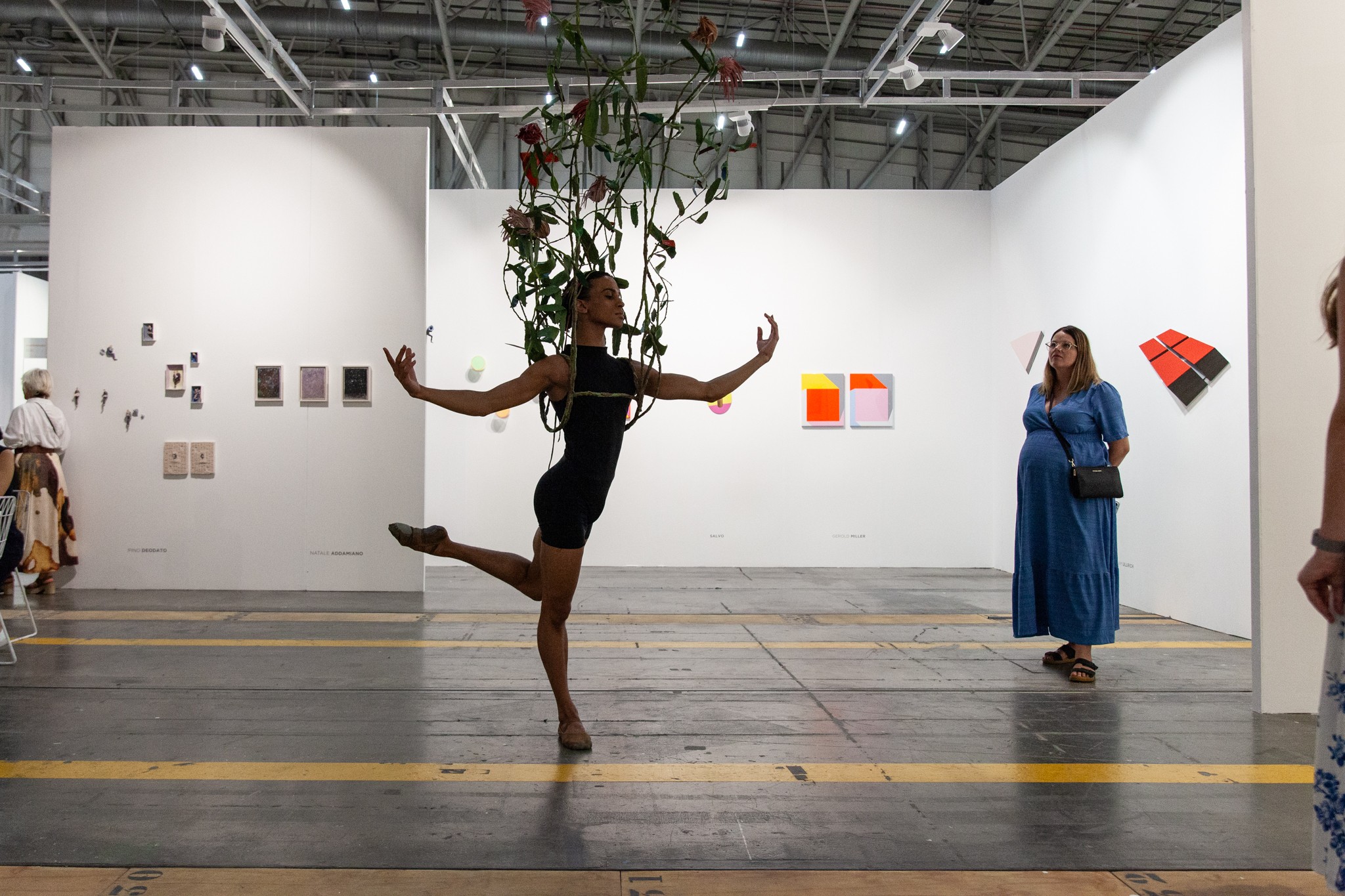 A dancer in a black leotard with long vines and red flowers extending from their back performs a ballet pose in a white-walled art gallery.