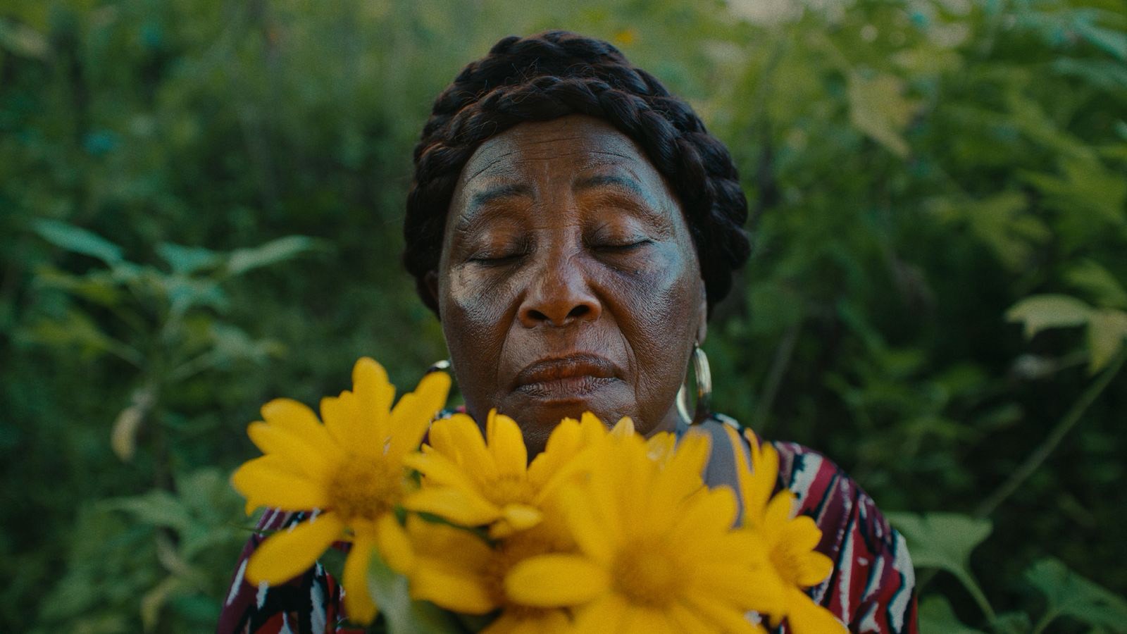 An older Black woman with braided hair and closed eyes holds yellow flowers.