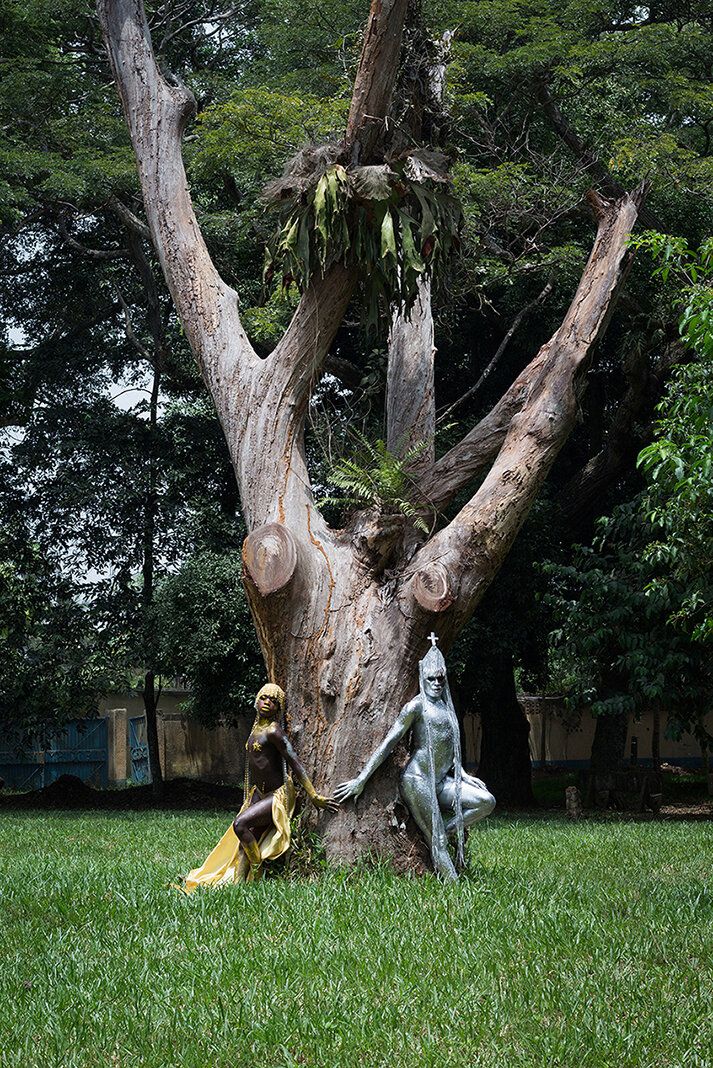 A gold-painted figure and a silver-painted figure with a mitre lean against a large, gnarled tree in a grassy park.