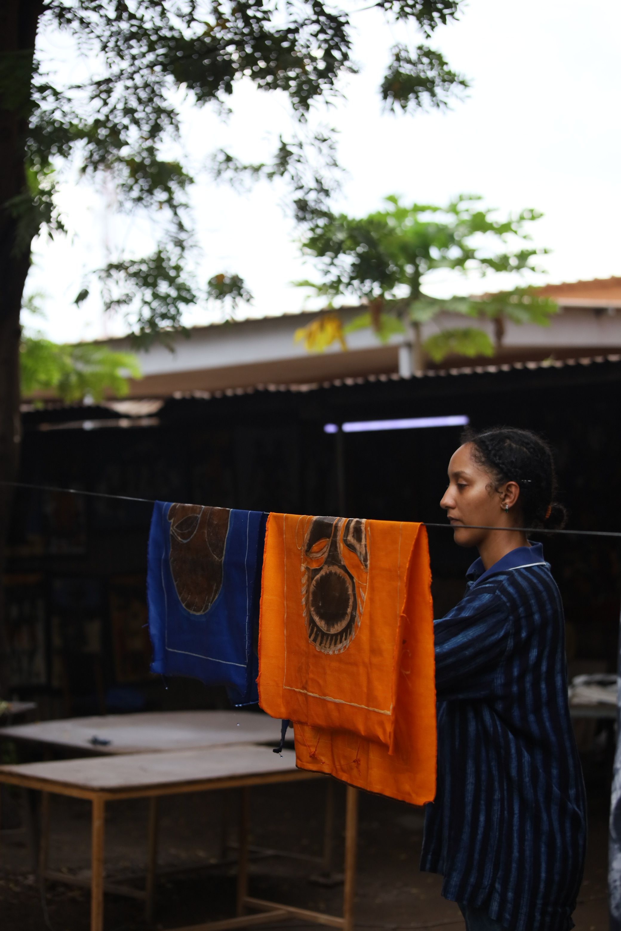 A young woman stands outdoors looking left, next to a line with blue and orange fabrics featuring dark mask-like designs.