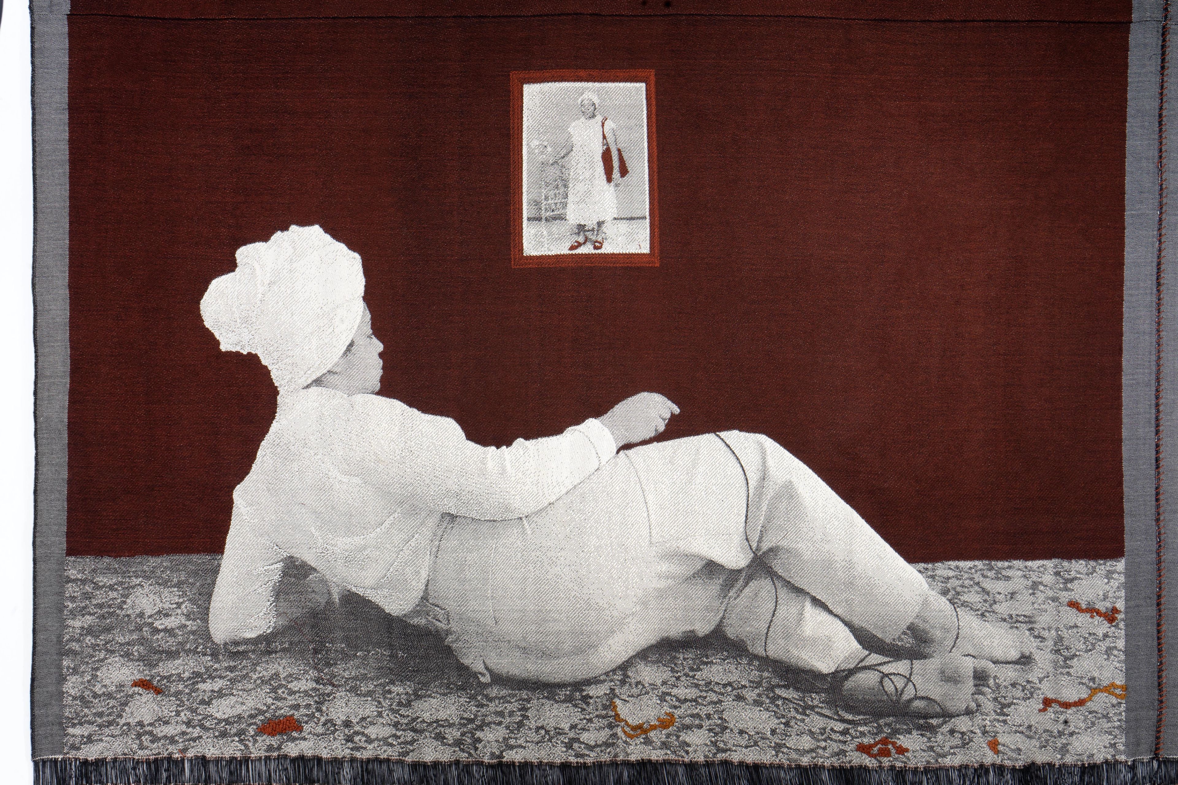 Tapestry depicting a reclining grayscale figure in white, wearing a turban, on a patterned rug, gazing at a framed grayscale portrait of a standing figure on a dark red wall.