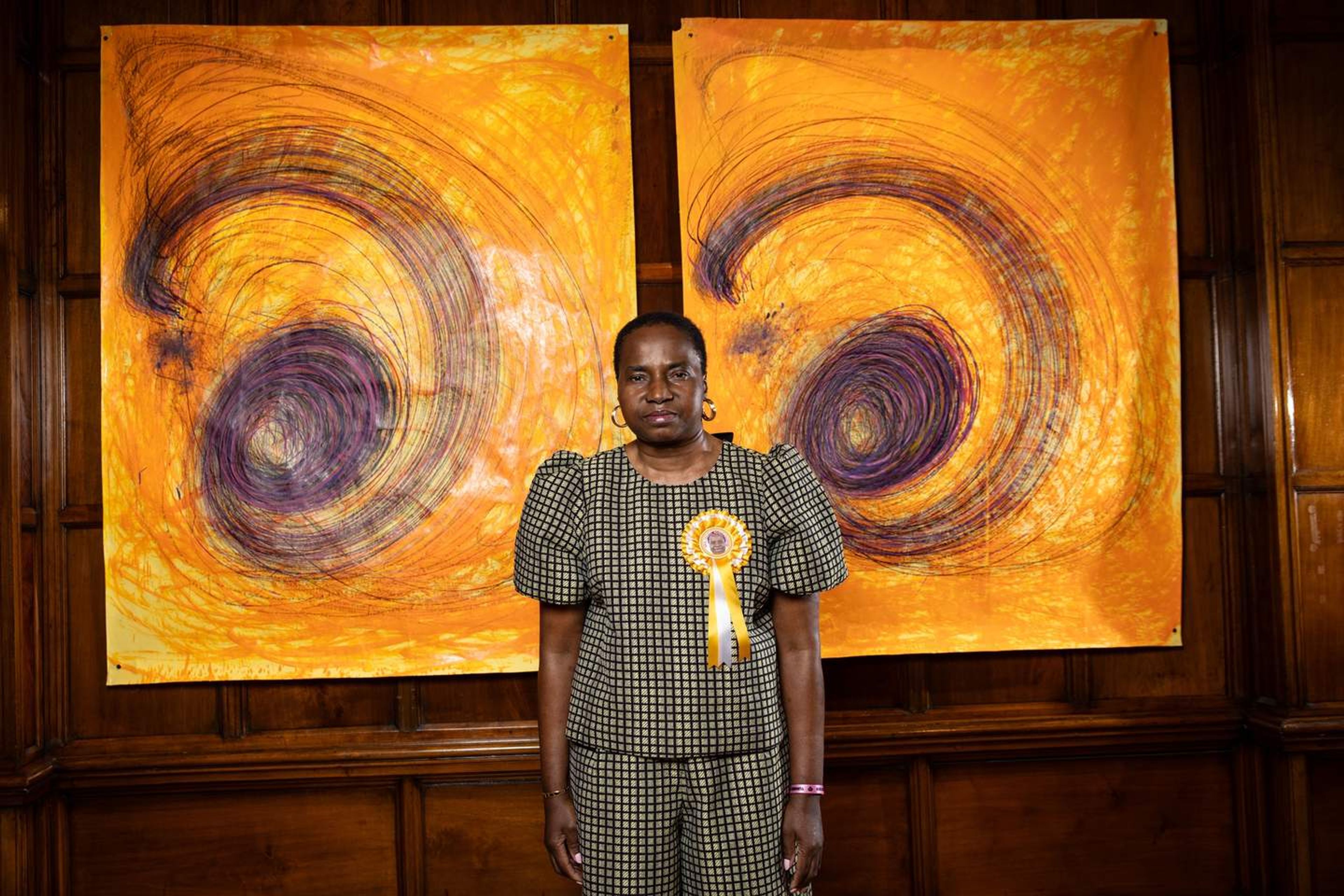 A Black woman wearing a patterned top and trousers, with a yellow rosette pinned to her chest, stands before two large, abstract orange and purple swirling paintings against a dark wooden wall.