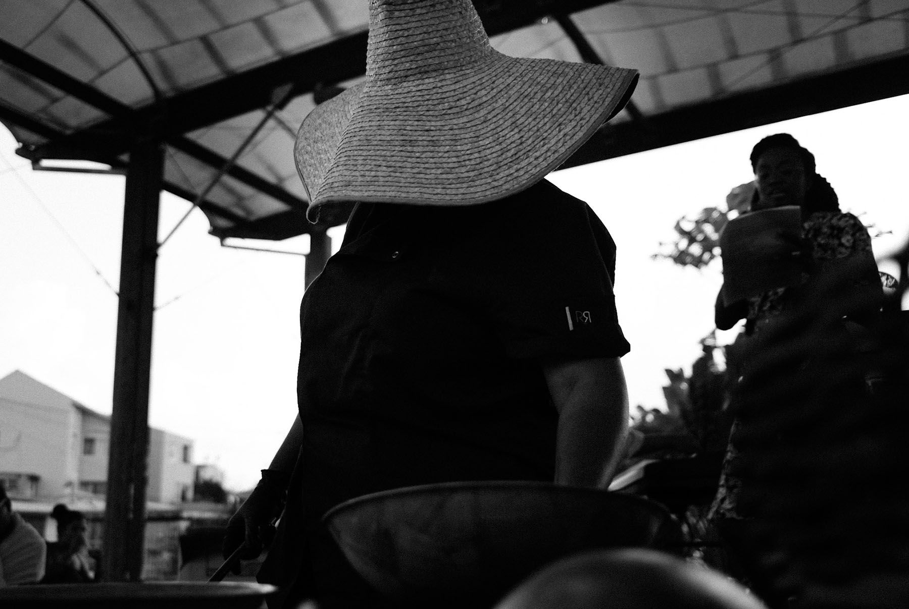 Black and white photo of a person wearing a wide straw hat that hides their face, standing at a market stall.
