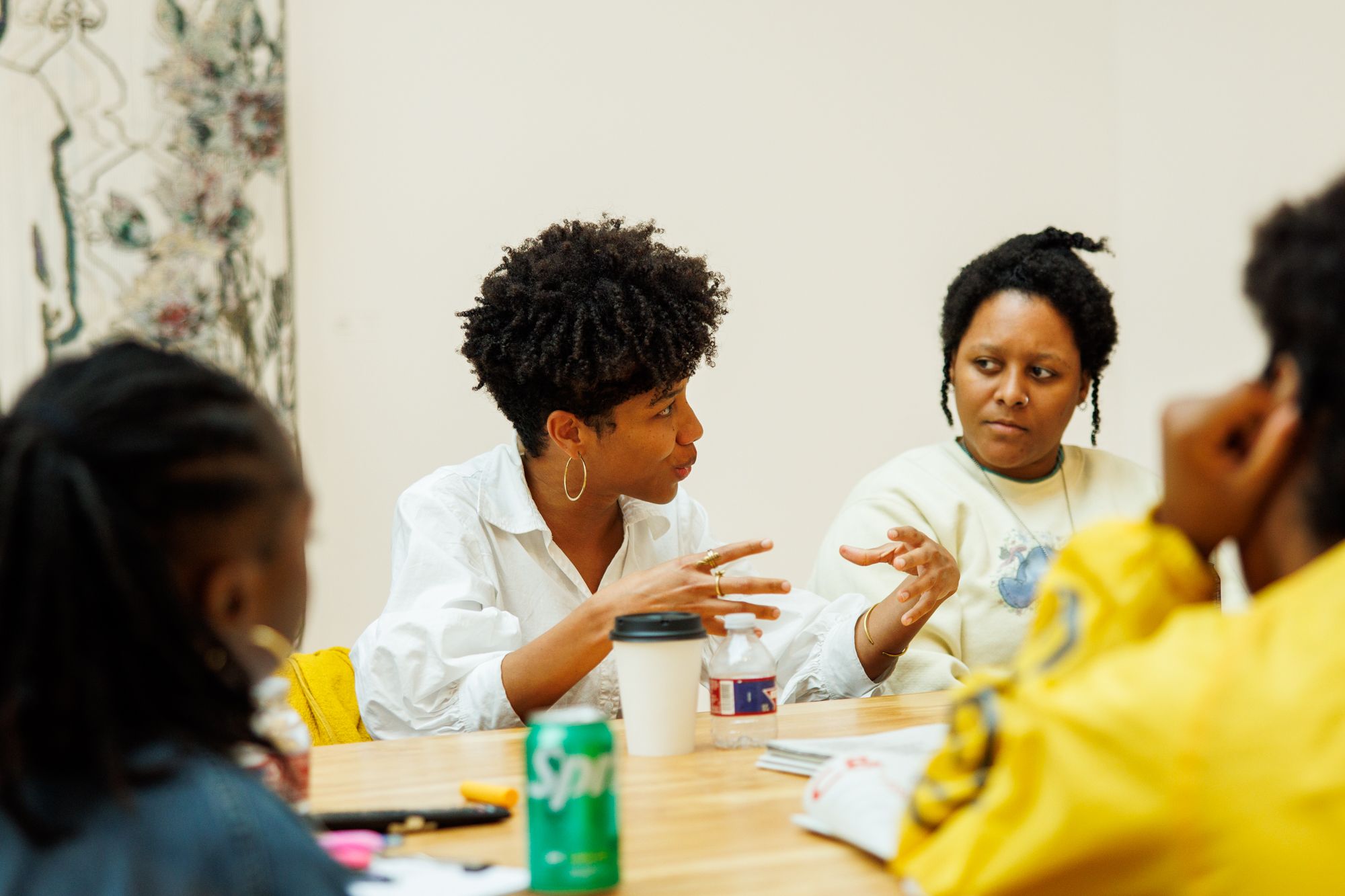 A Black woman with short curly hair speaks animatedly to another woman across a table, with two other people partially visible.