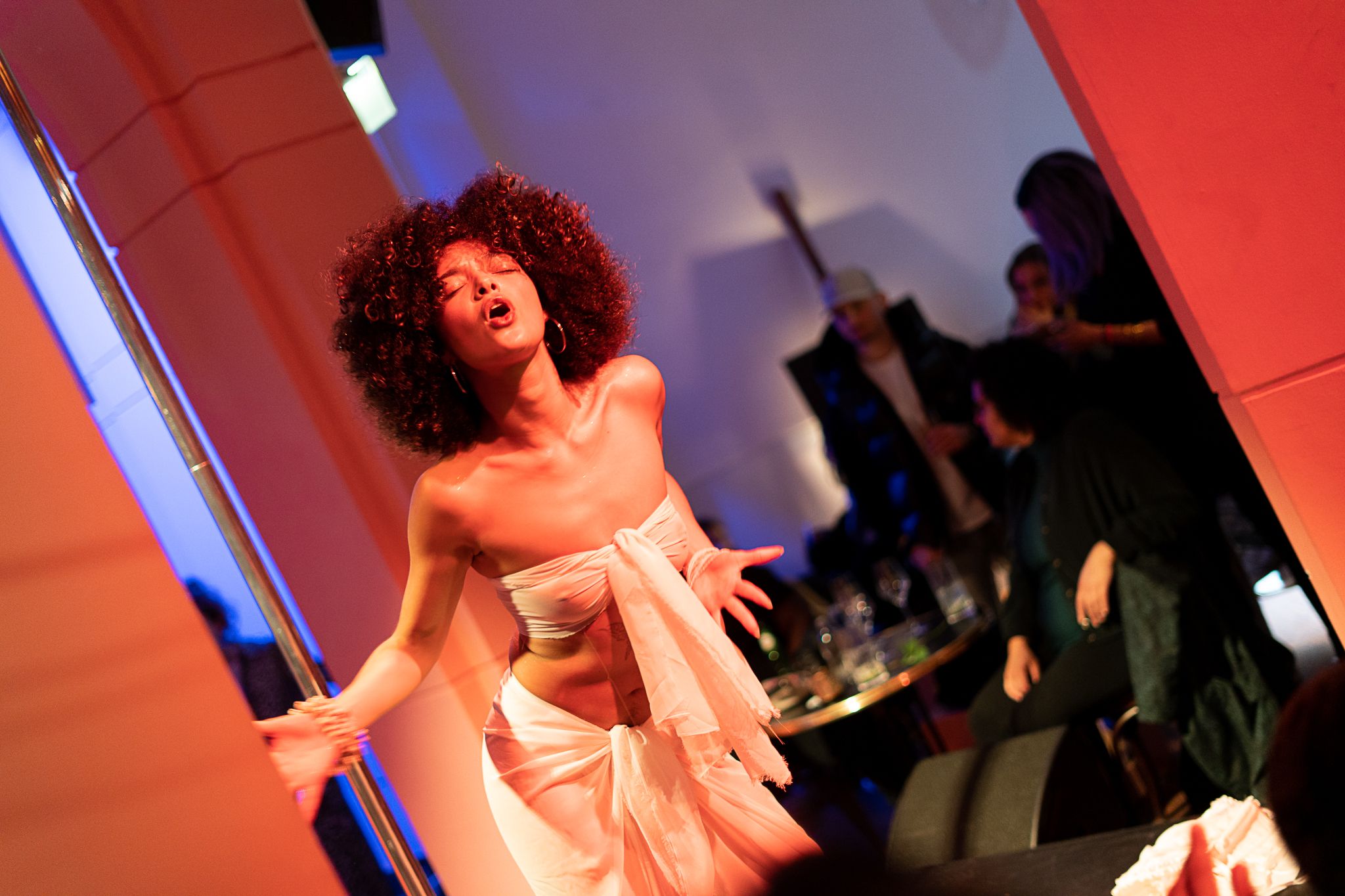 A woman with voluminous curly hair, wearing a white draped outfit, sings passionately under orange and blue stage lights, with blurred figures in the background.