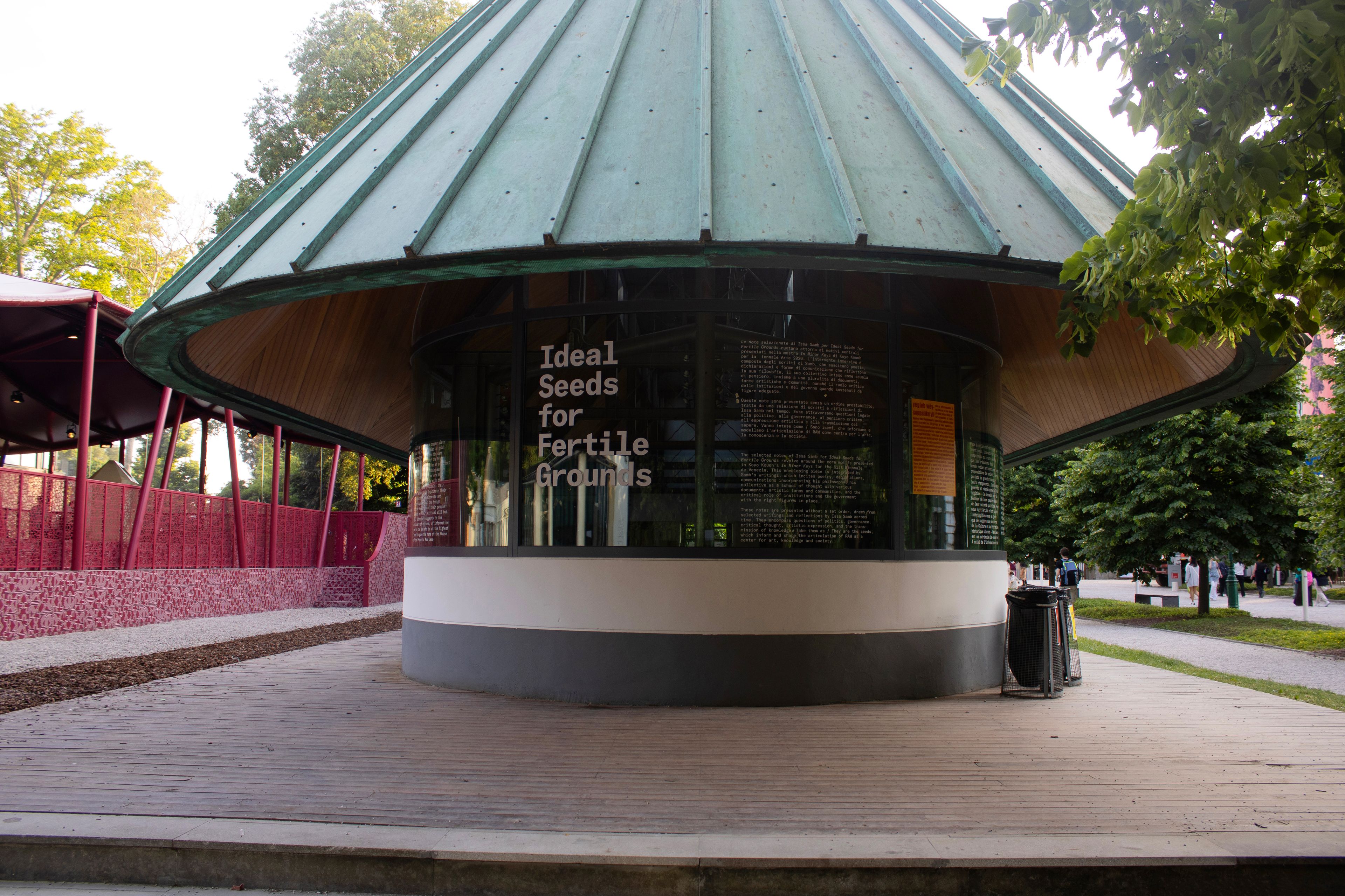 A circular pavilion with a green roof and glass walls displaying "Ideal Seeds for Fertile Grounds" on a wooden deck in a park.